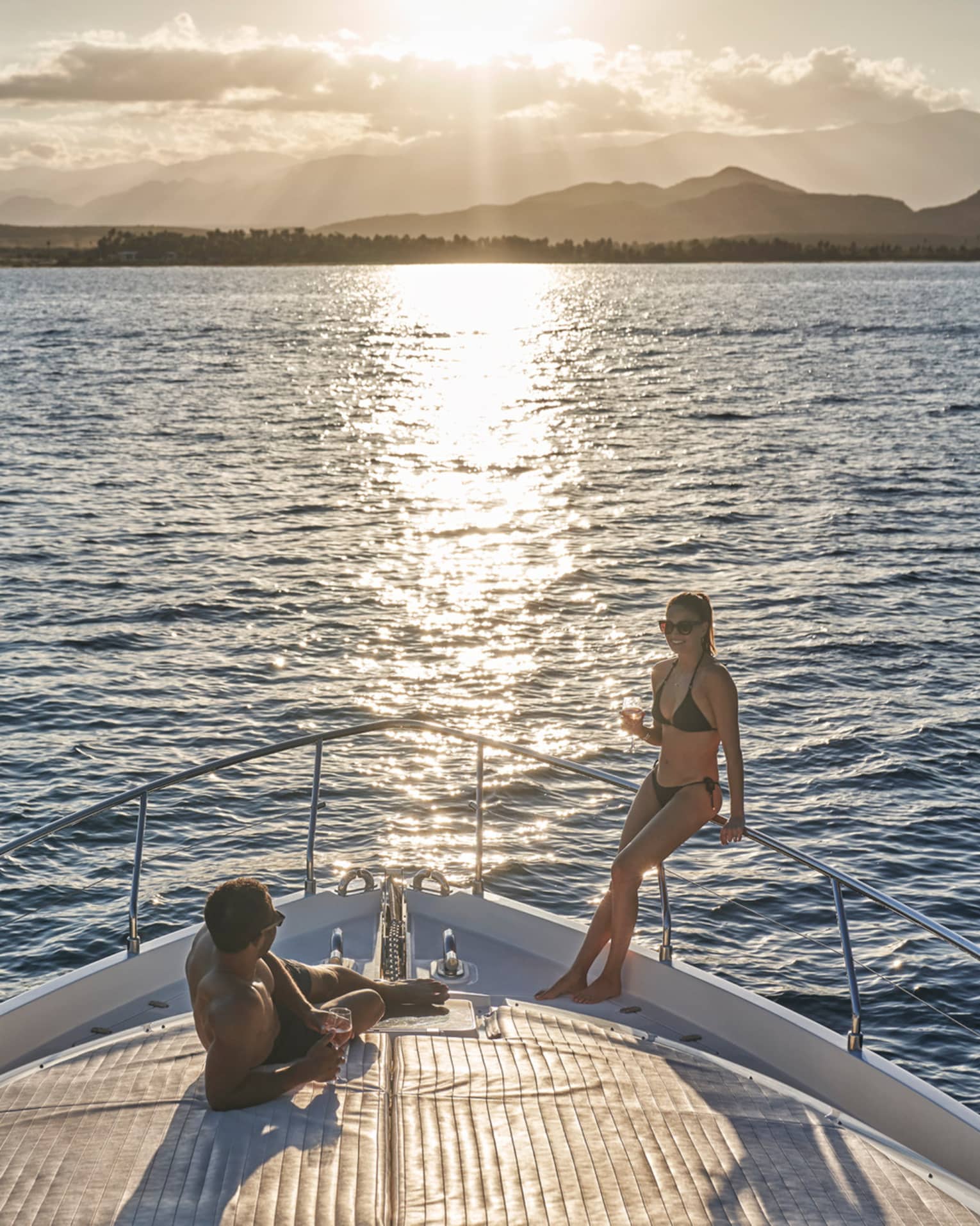 A couple in swimwear relaxing on the bow of a boat, wine glasses in hand and the setting sun glinting off the endless ocean.
