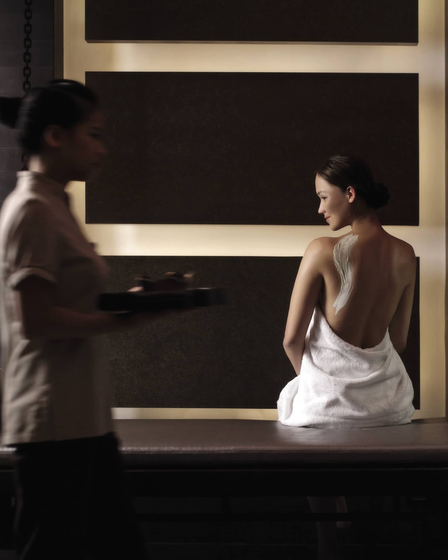 Woman in white towel with cream on back sits on bench in dark spa room