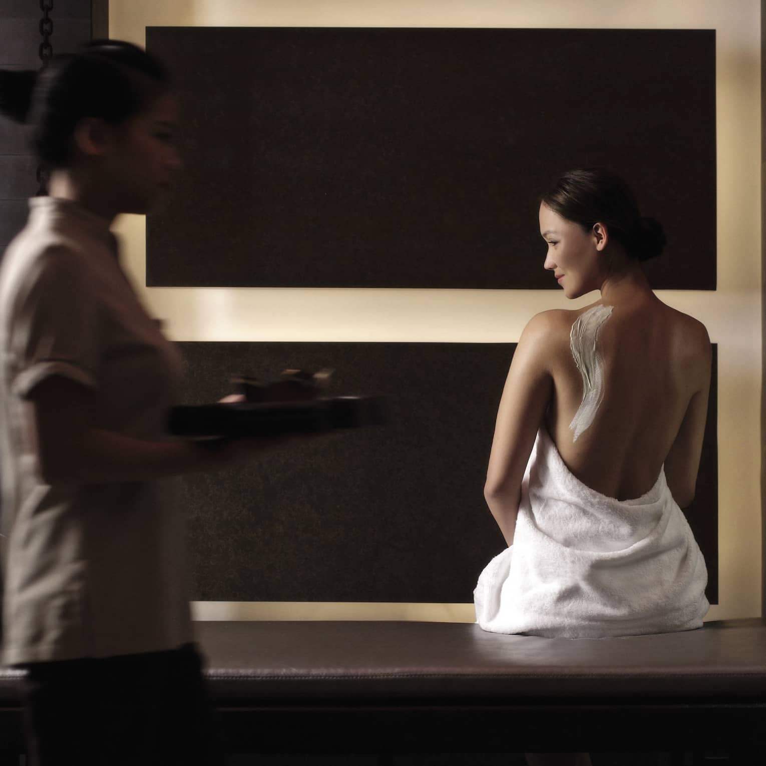 Woman in white towel with cream on back sits on bench in dark spa room