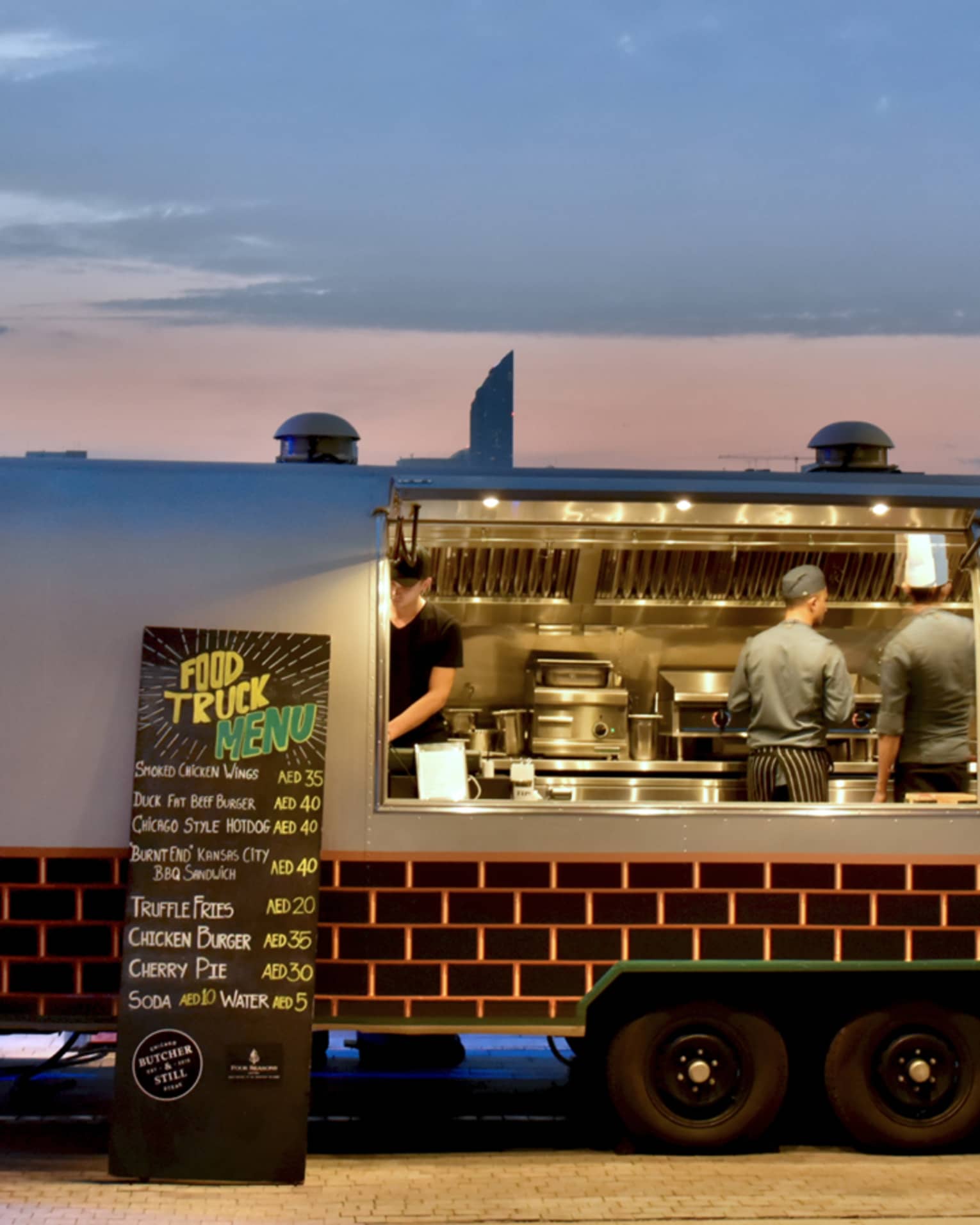 The Butcher and Still airstream food truck is open for business with the door opened, chefs cooking, and sun setting in the distance 