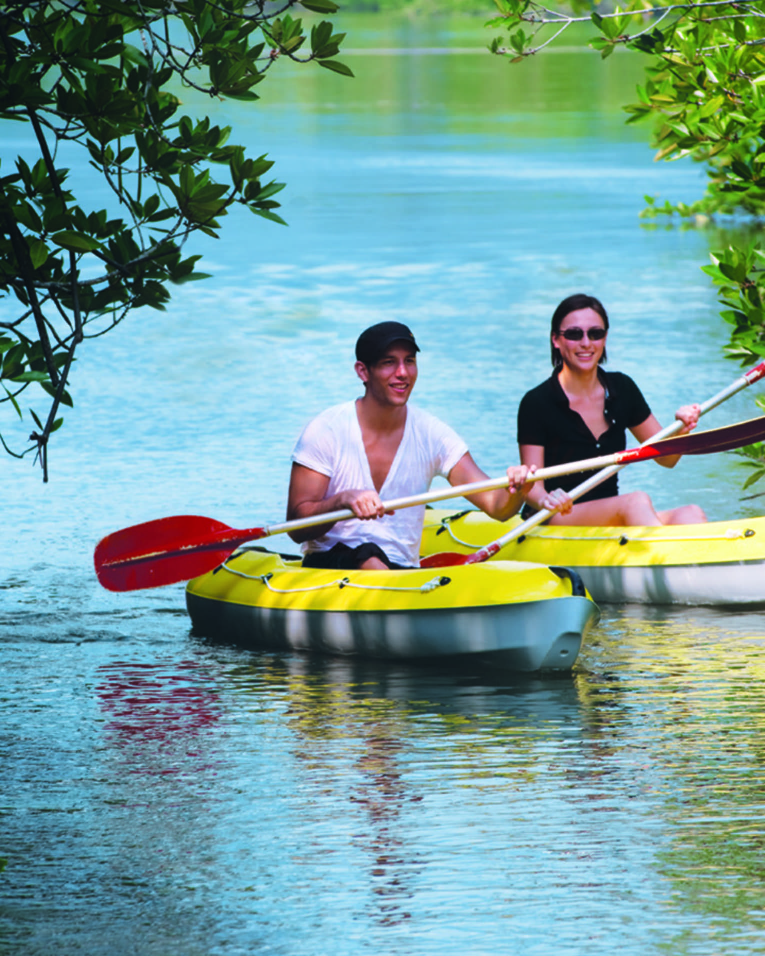 Two people with paddles in yellow kayaks pass through trees on water 