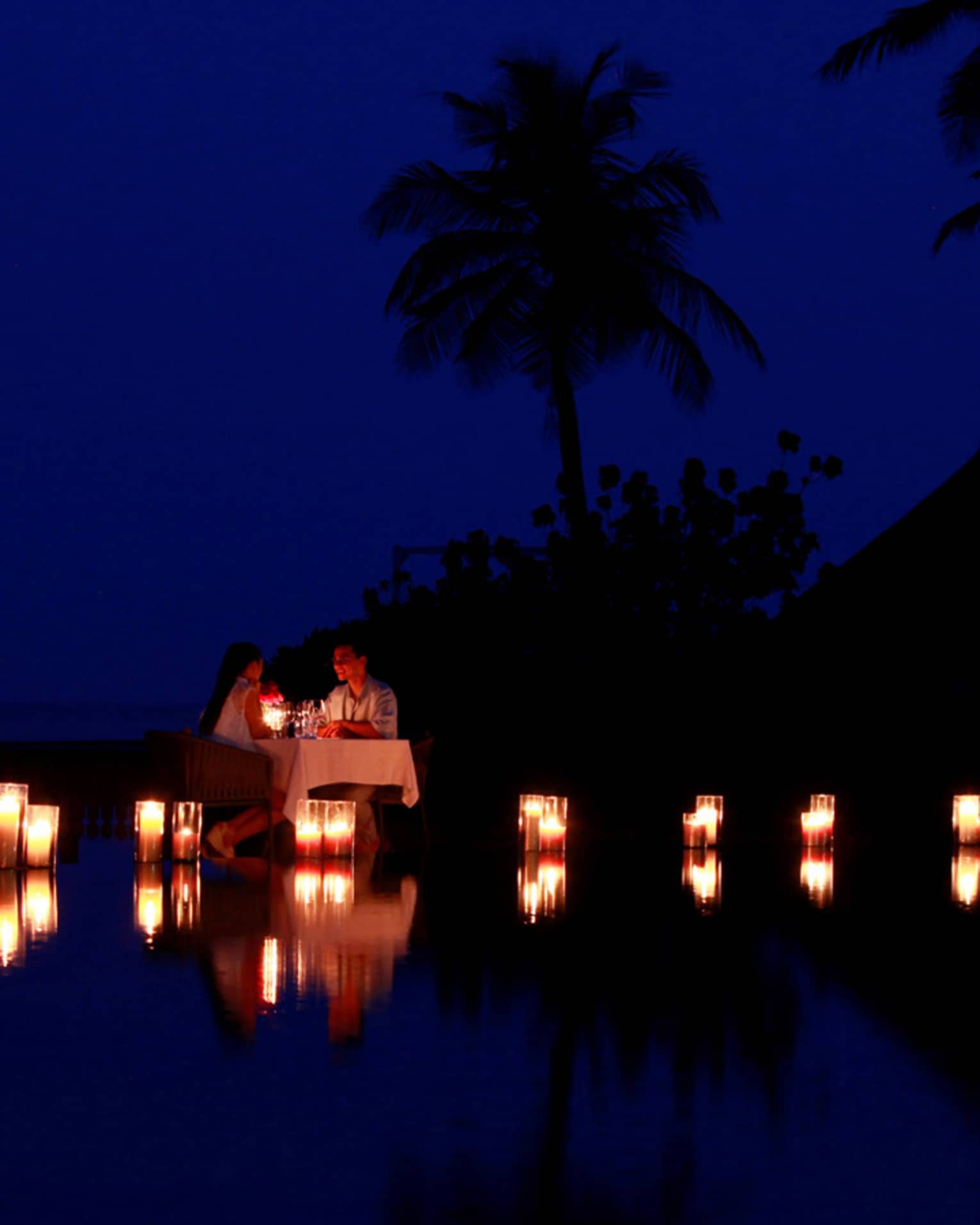 Couple dine by lagoon at small table surrounded by glowing candles at night