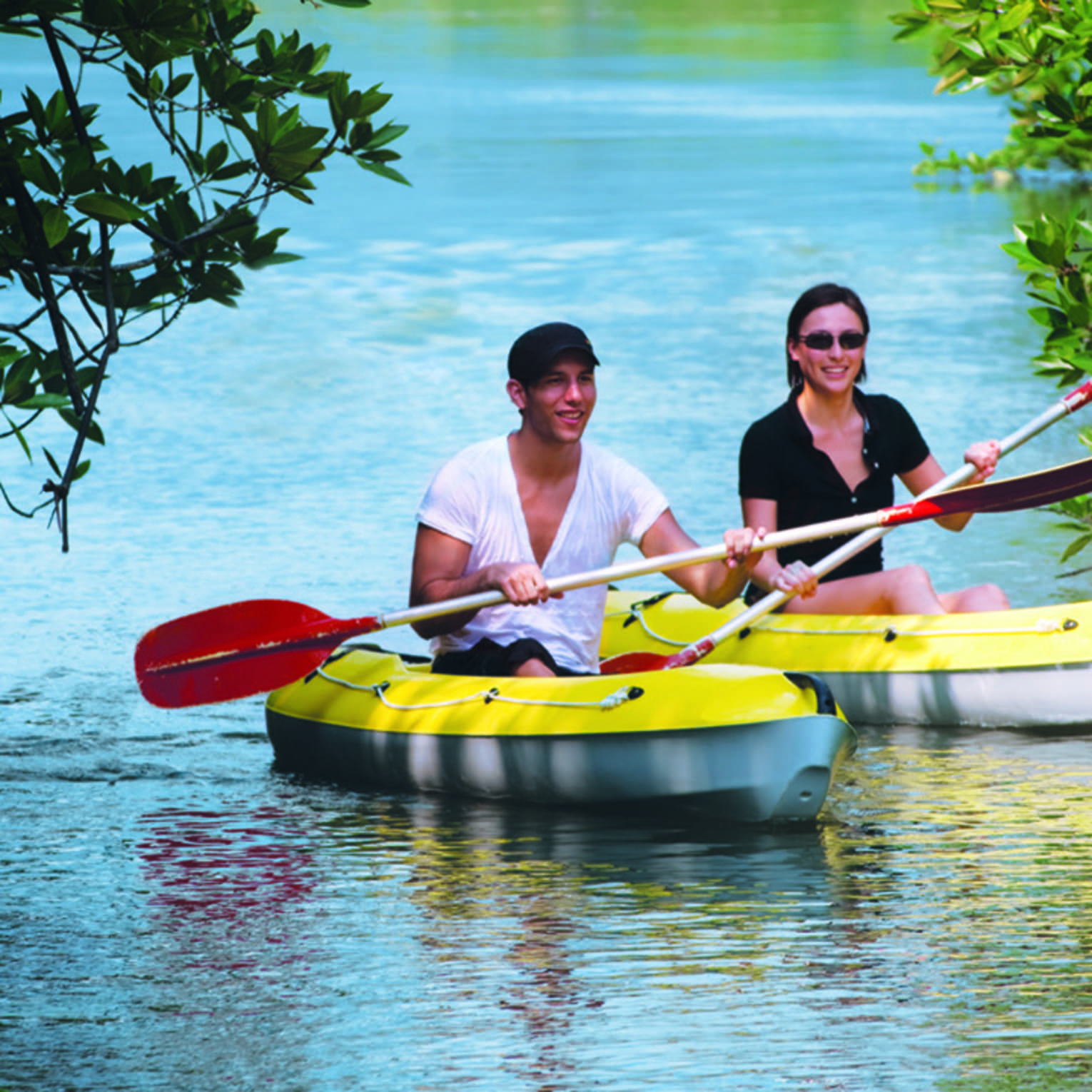 Two people with paddles in yellow kayaks pass through trees on water 