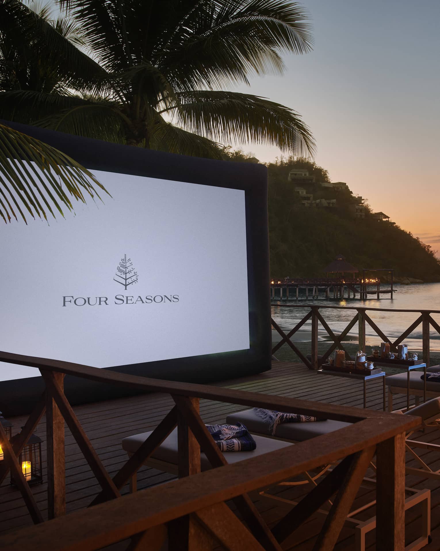 Under a dusky pink sky near calm water, lounge chairs face a large movie screen on a wooden dock covered by lit lanterns.