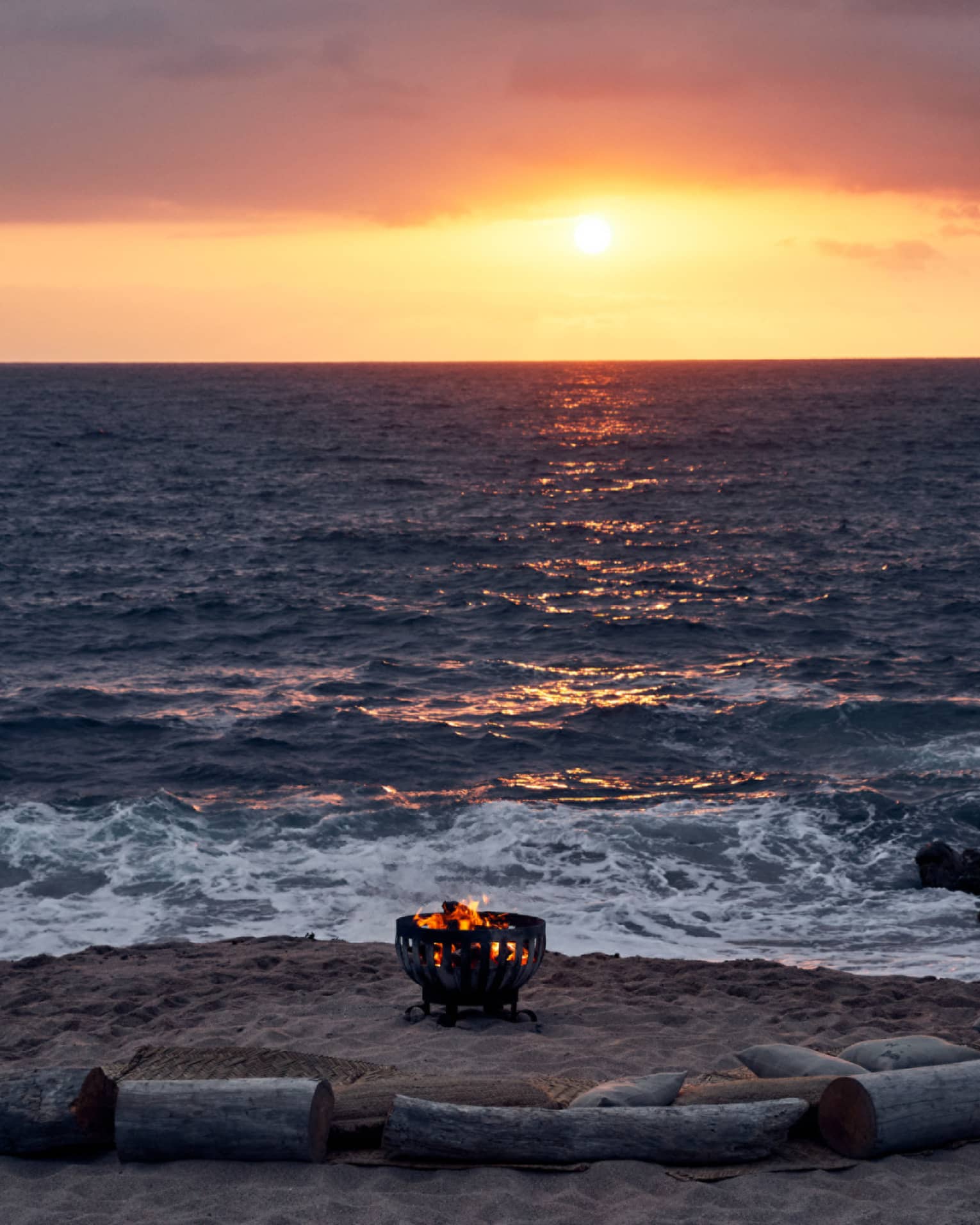 Fire burns in an iron pit on the beach, semi-encircled by driftwood log seats facing the sun setting into the ocean horizon.