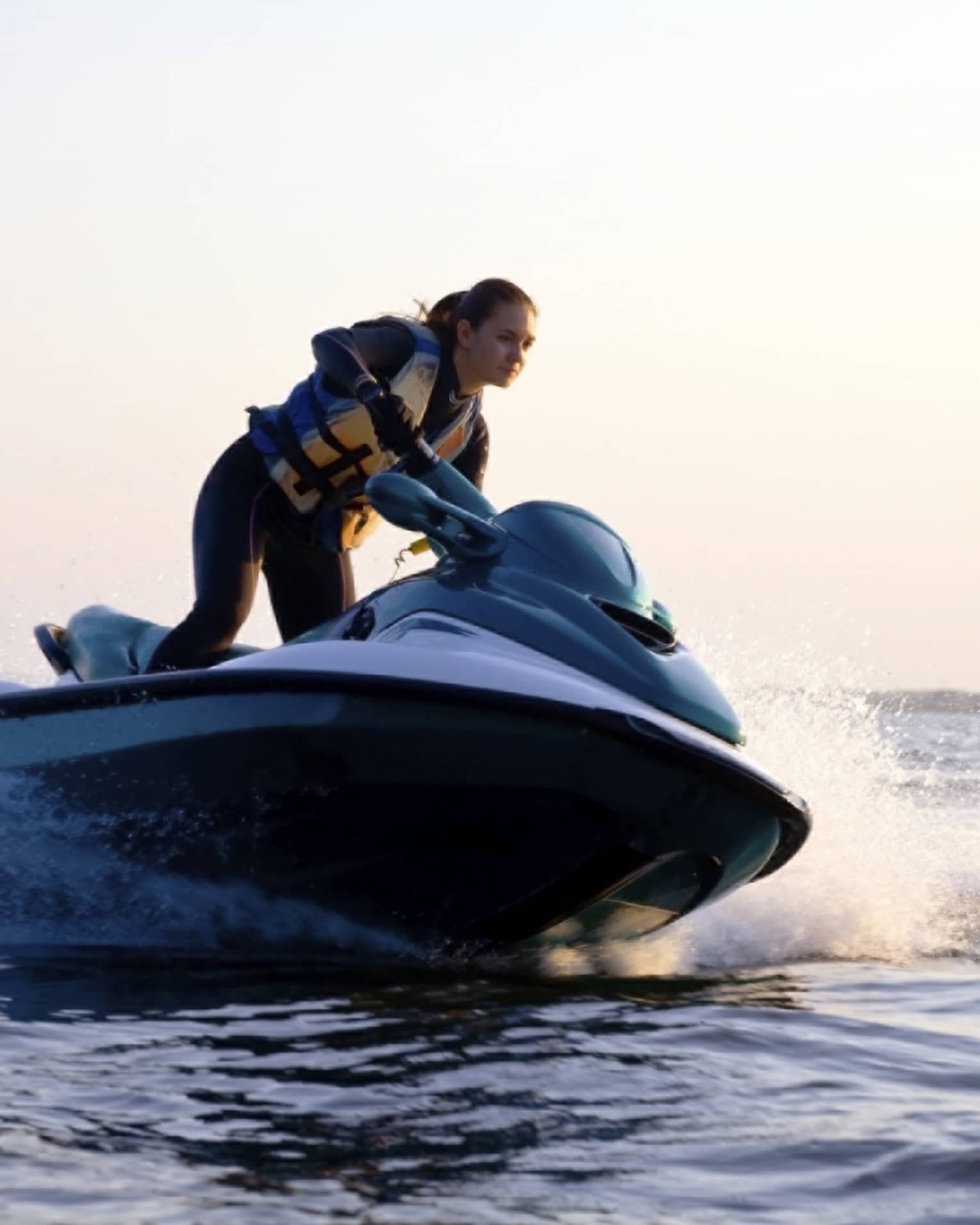 Woman wearing life jacket stands, steers jet ski on ocean at sunset