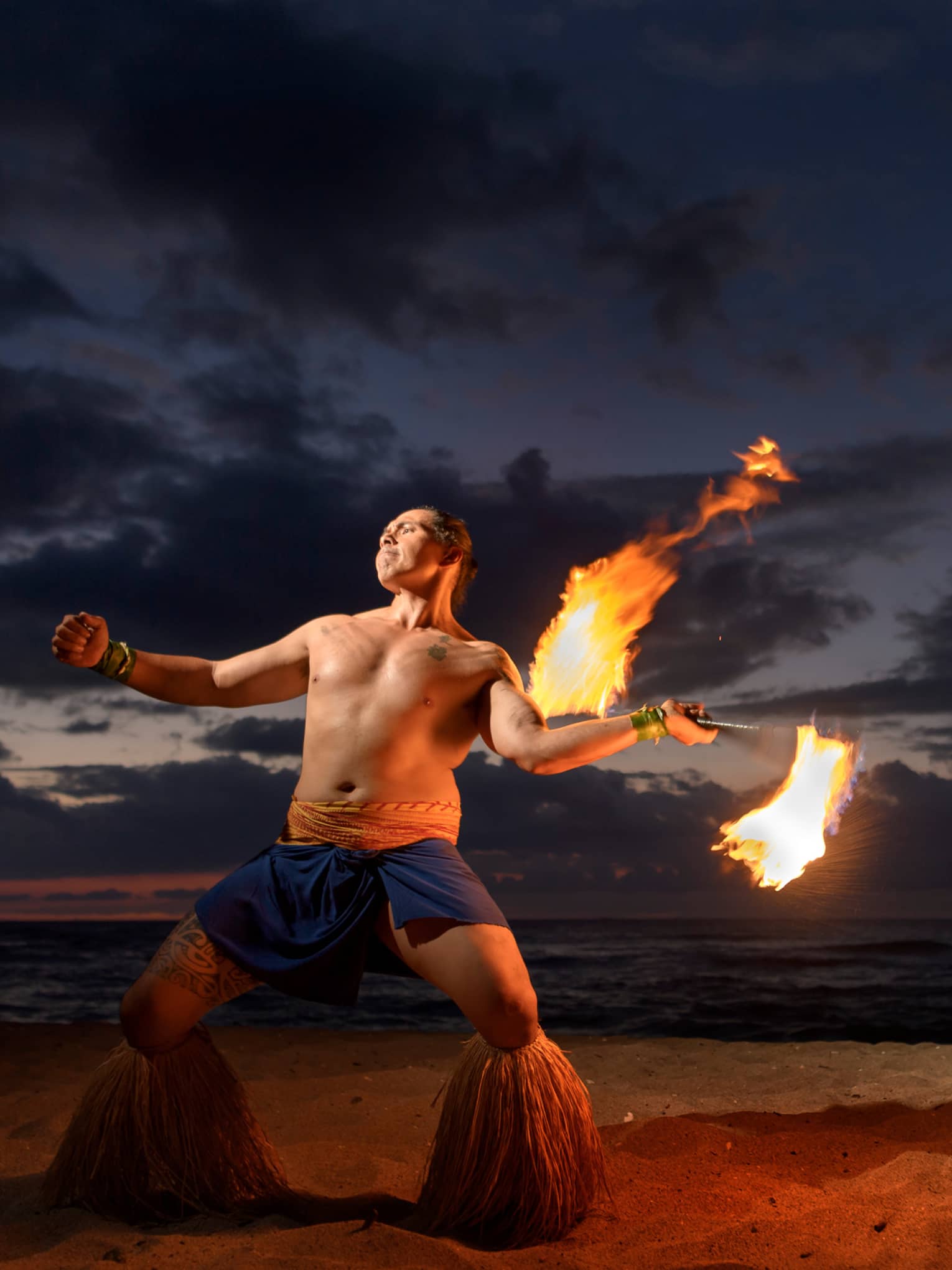Man wearing traditional Polynesian outfit dances with torch on beach at night
