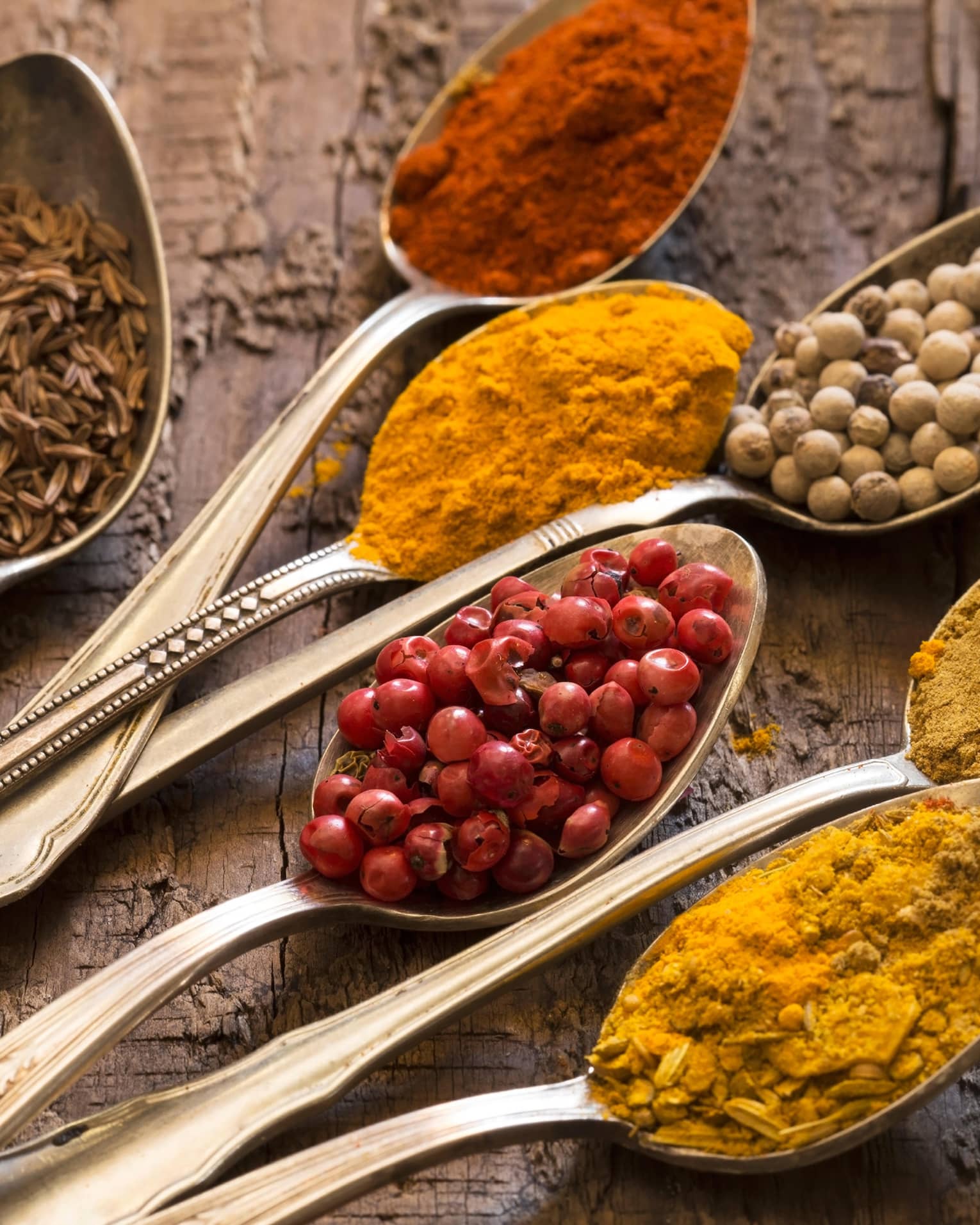 Seven silver spoons filled with various spices.