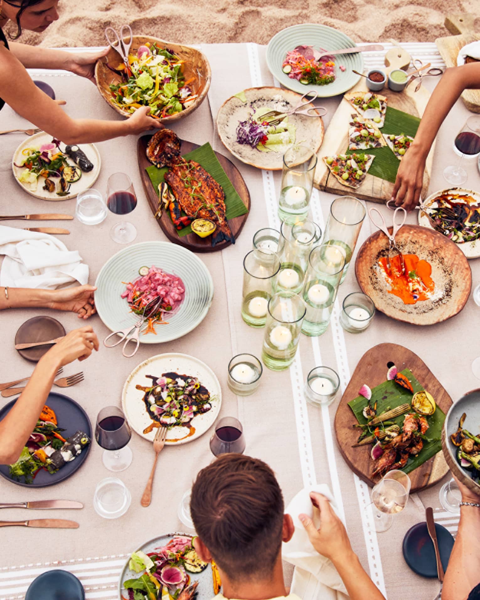 Beachside dining table with an assortment of vibrant dishes, candlelit glass holders and guests enjoying fresh seafood and grilled vegetables