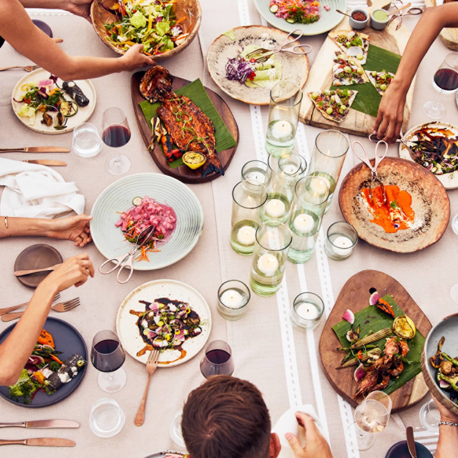 Beachside dining table with an assortment of vibrant dishes, candlelit glass holders and guests enjoying fresh seafood and grilled vegetables