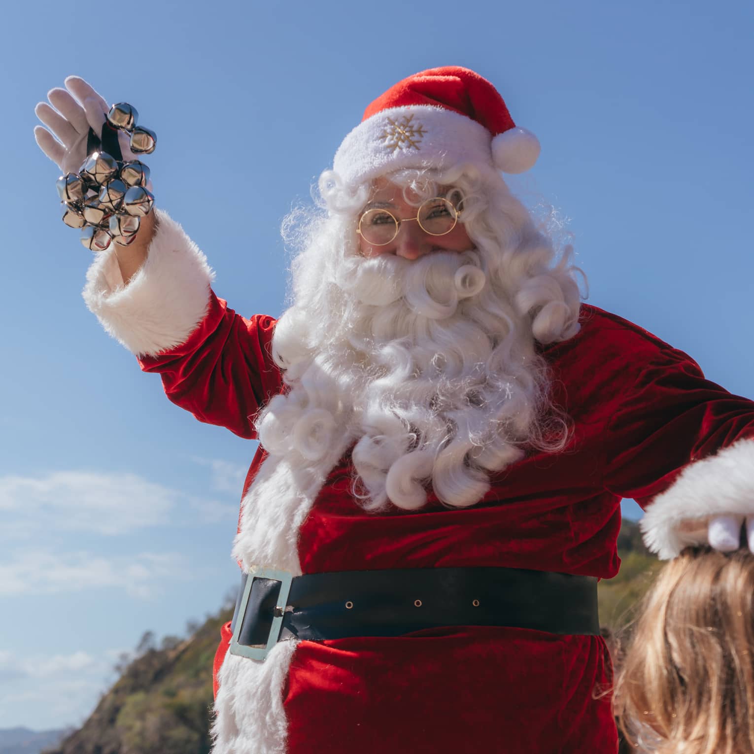 Person dressed in a red Santa Claus suit and white beard holds jingle bells in their hand and waves while standin next to a child on a beach