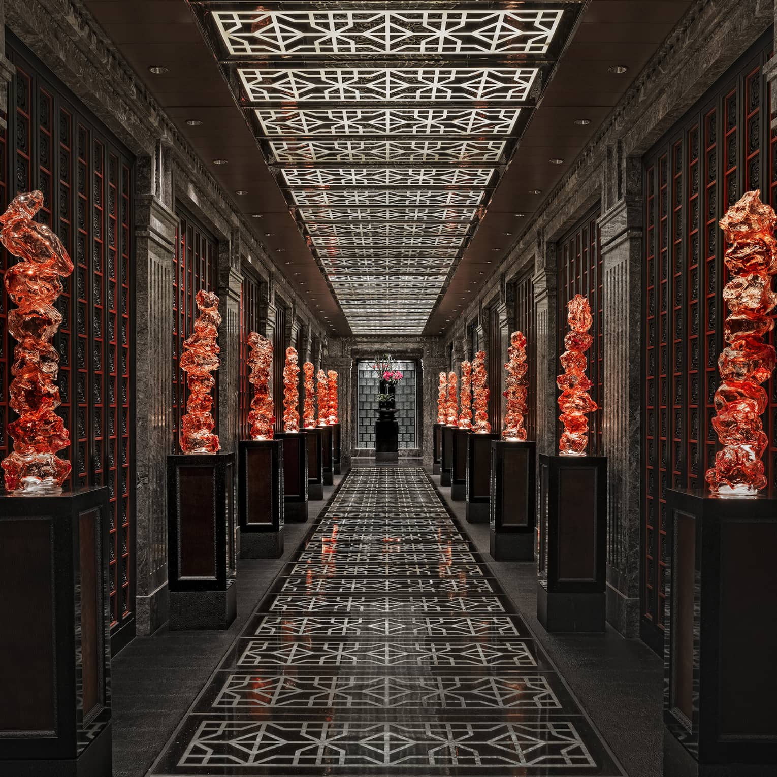 View down a long black-tiled hall lined with orange glass sculptures on pedestals, the geometric ceiling mirroring the floor.