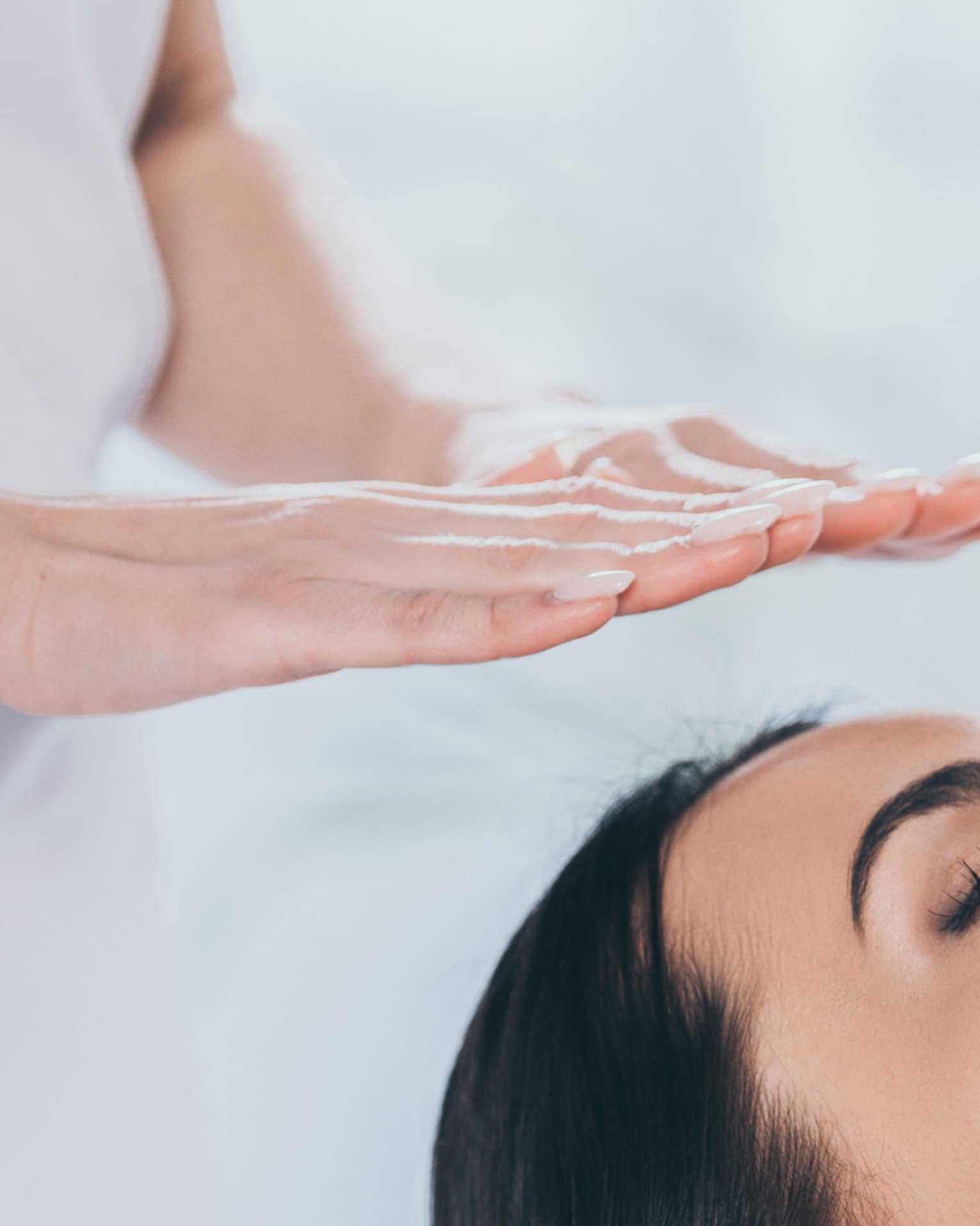 Woman with eyes closed lays on rolled up towel on spa table under therapists' hovering hands