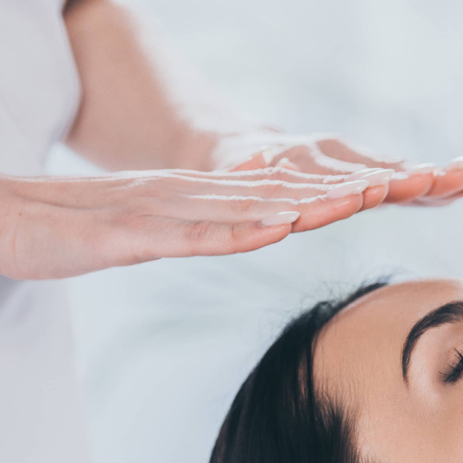 Woman with eyes closed lays on rolled up towel on spa table under therapists' hovering hands