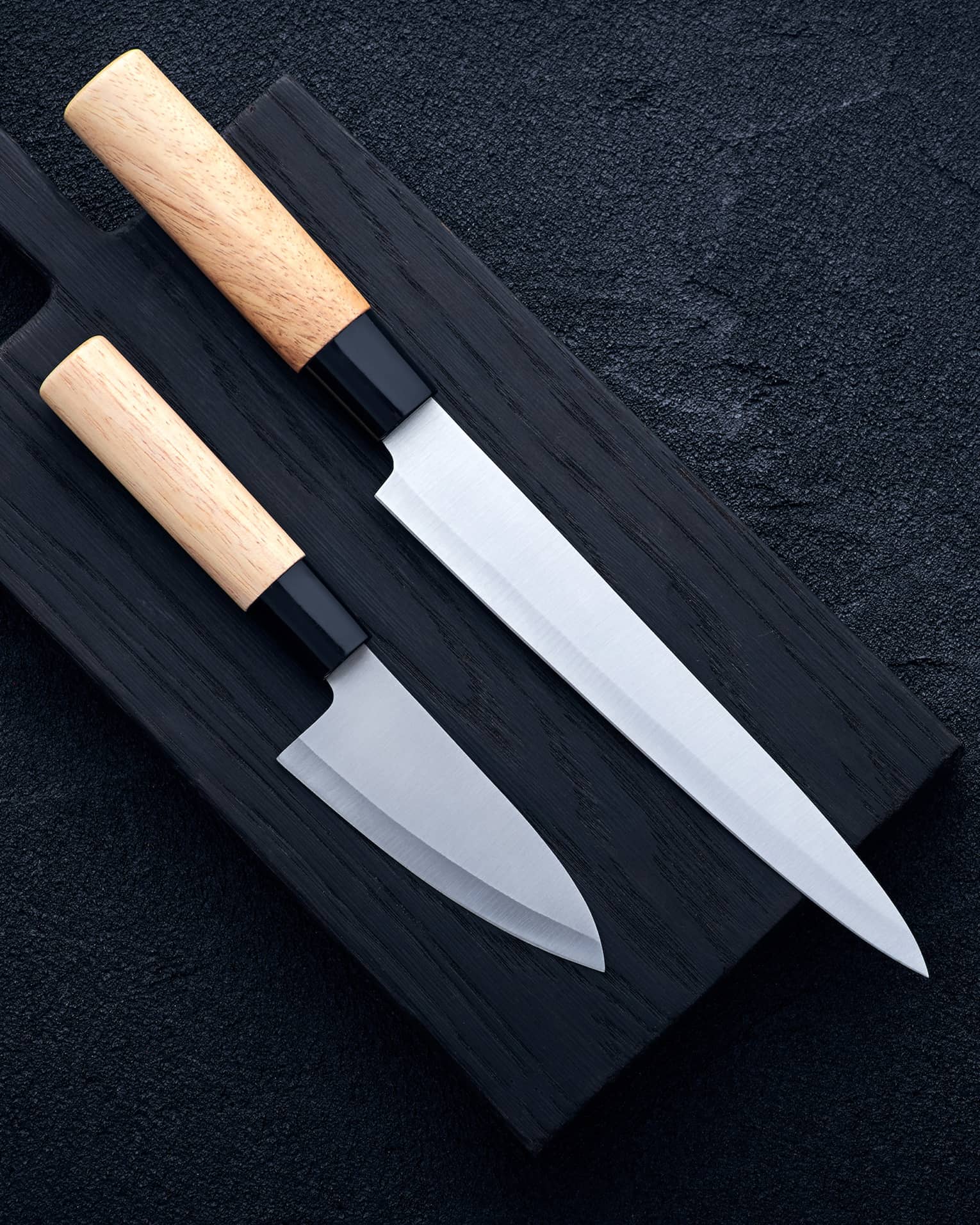 Two extremely sharp wood-handled knives, one long, one short, laid out on a black cutting board on a dark surface.