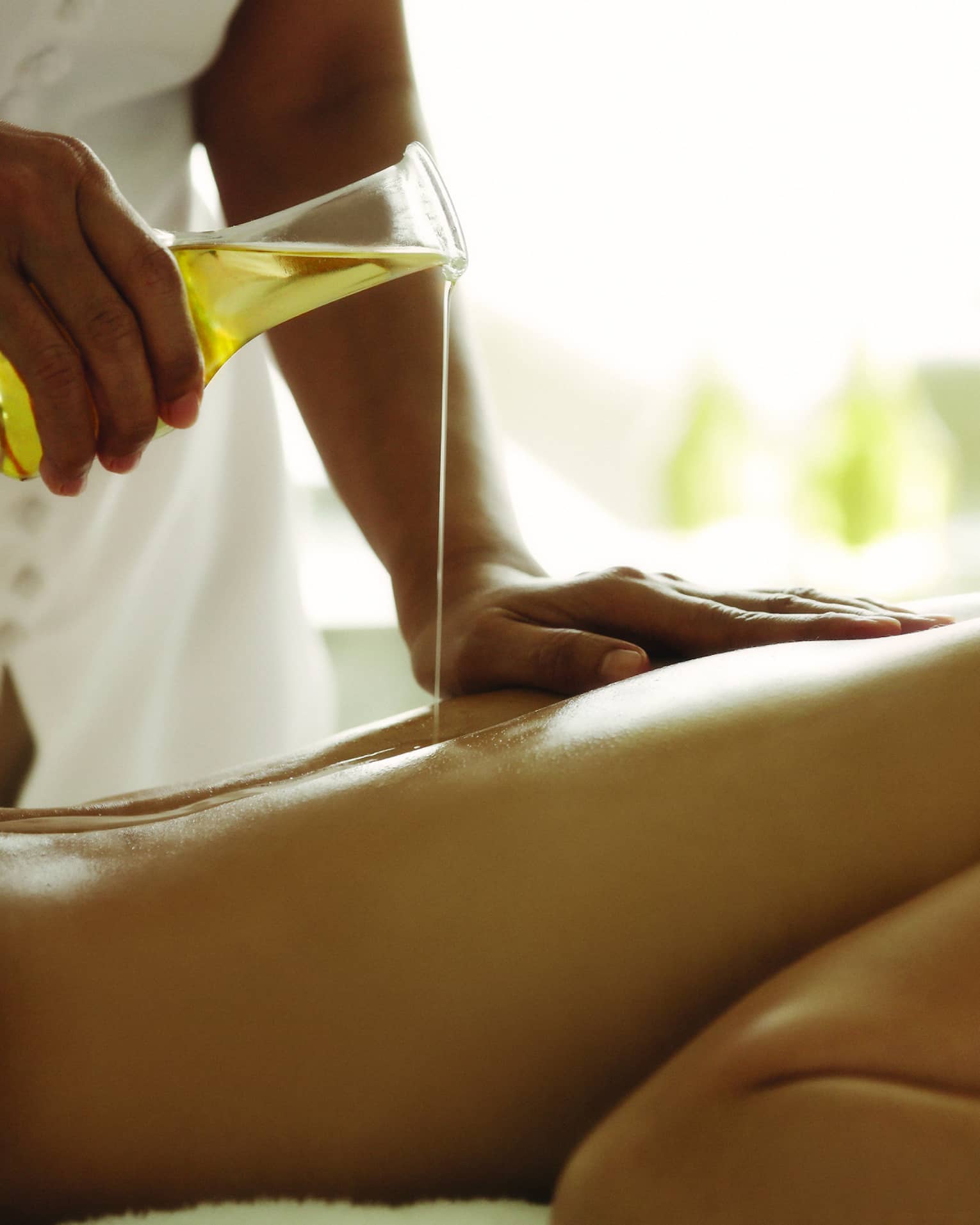 Spa staff pours massage oil over woman's bare back as she lays on treatment bed