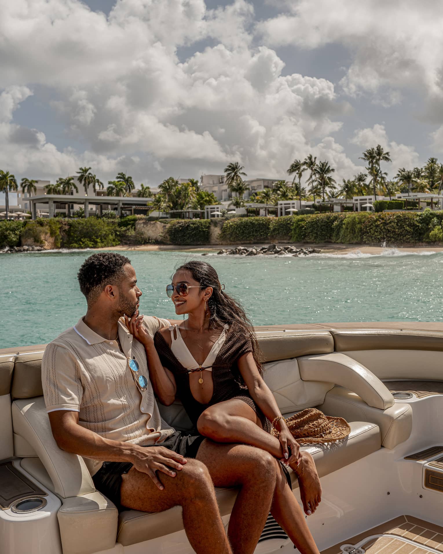 Two people sit on luxury yacht at sea, with palm trees and resort in the background
