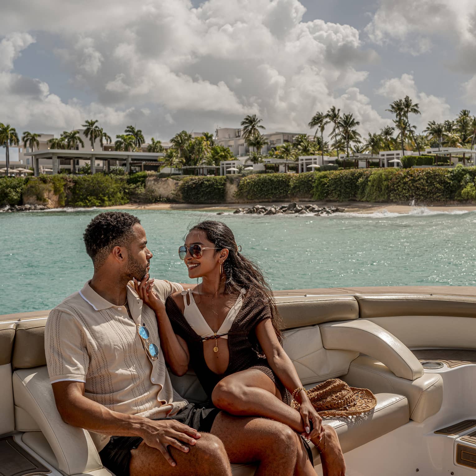 Two people sit on luxury yacht at sea, with palm trees and resort in the background