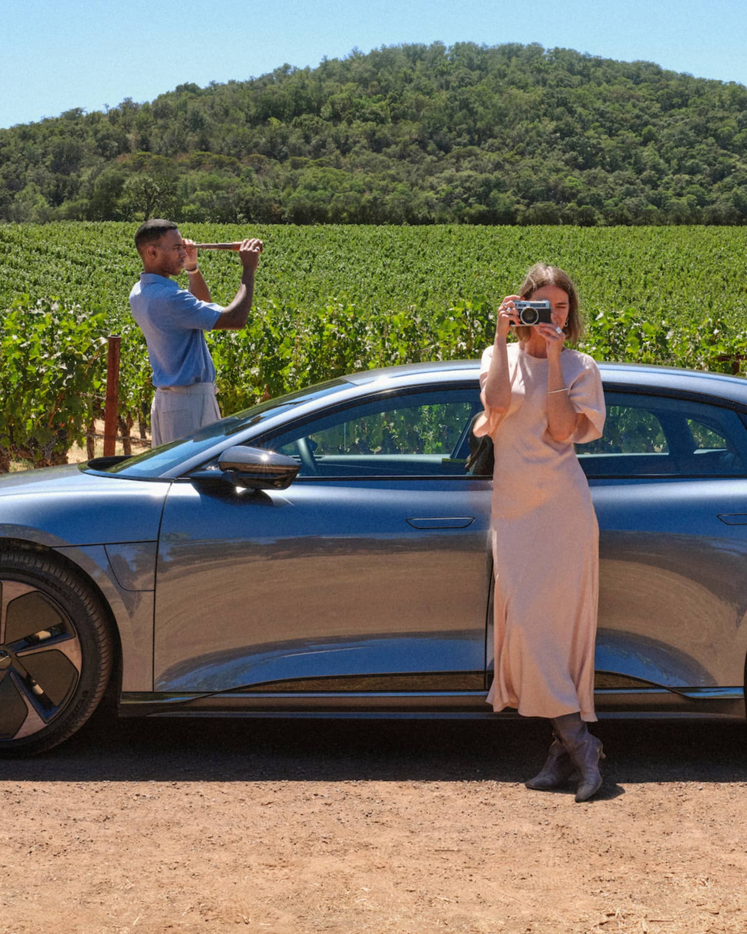 Person wearing long light-pink dress stands leaning against a grey car taking a picture of the surrounding vineyards with their camera while another person wearing a blue shirt and white pants stand on the other side of the car