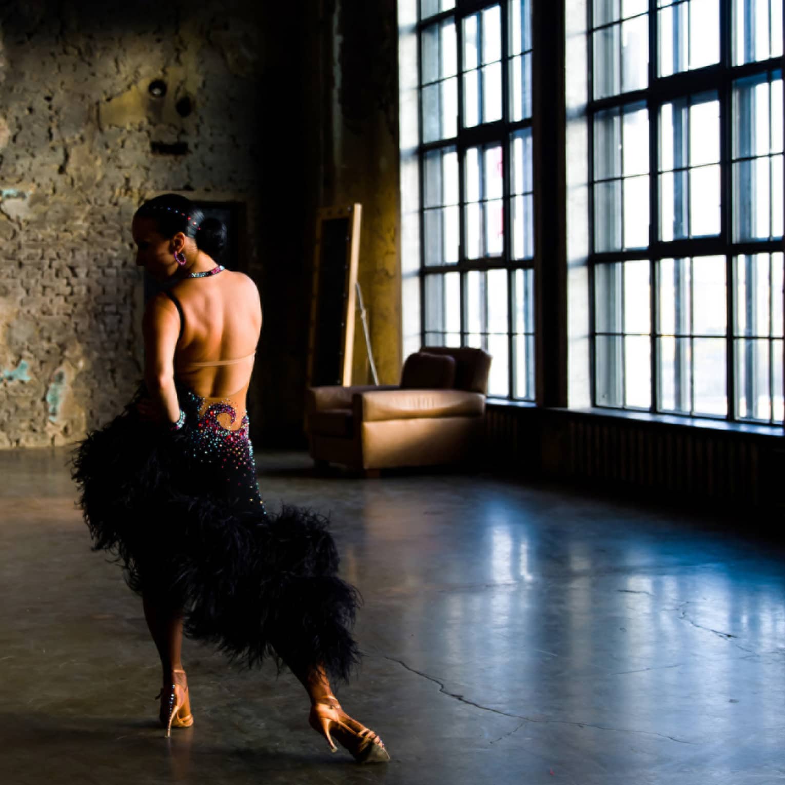 In an unlit room with large windows, a salsa dancer in an elaborate costume poses, hand on hip, opposite a brick wall. 