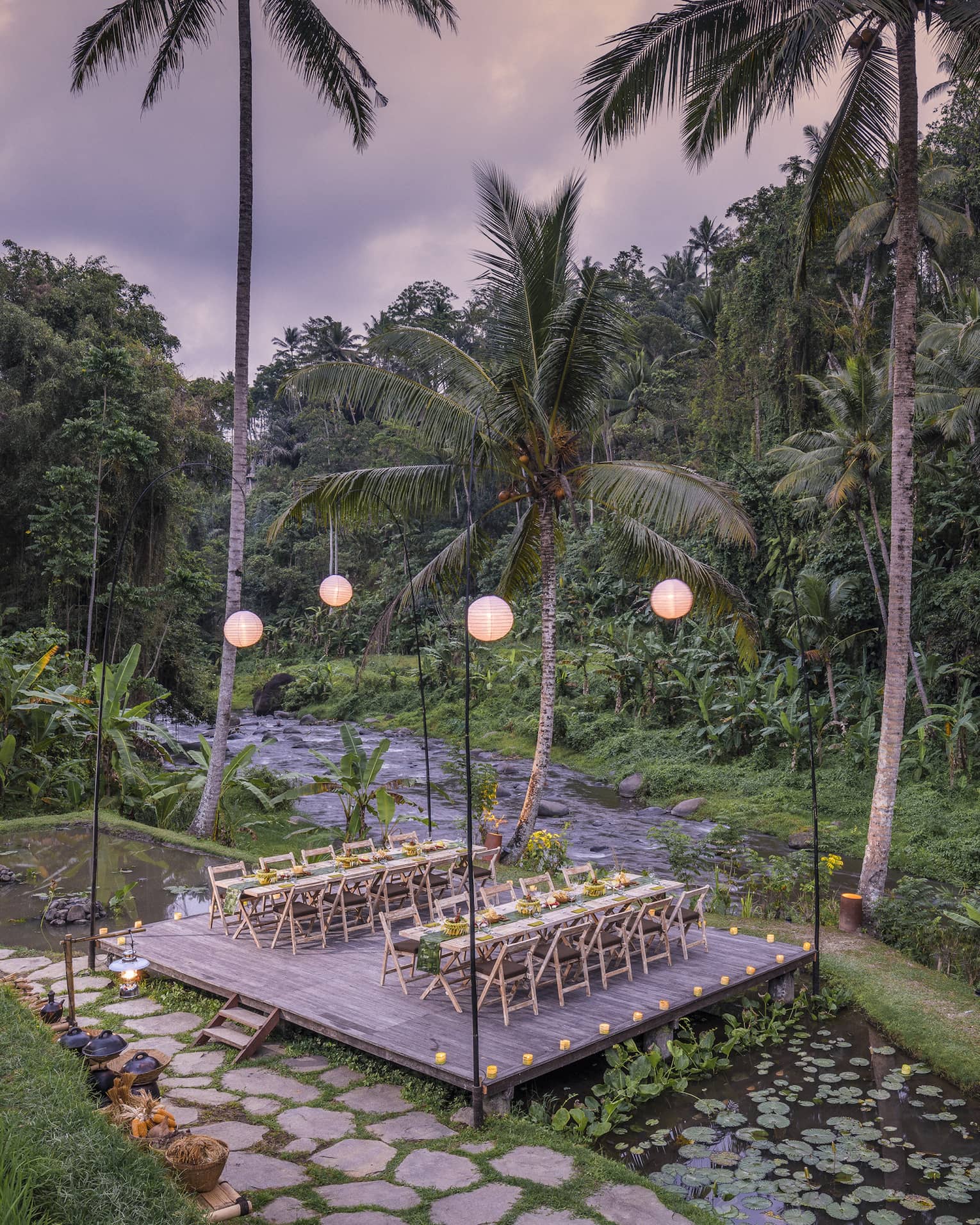 Two long tables laid out with food sit on a lantern-lit platform above a lotus pond and beside a river and dense forest.