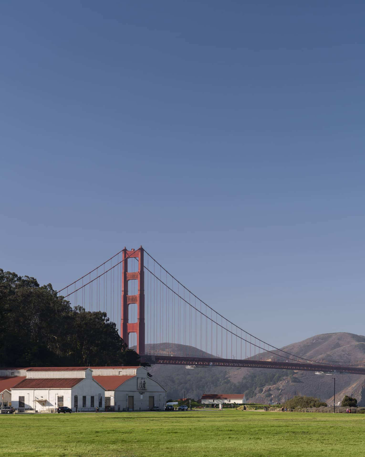 Red-roofed white buildings at the far end of a green field with the Golden Gate Bridge, rolling hills and a blue sky beyond.