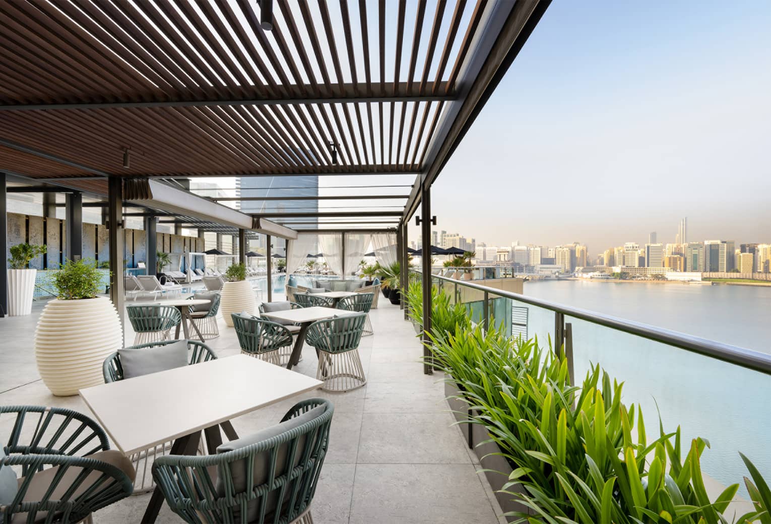 Restaurant terrace with slated wood overhang, various white tables, dark-teal chairs and a row of green plants overlooks the water with a city skyline in the background
