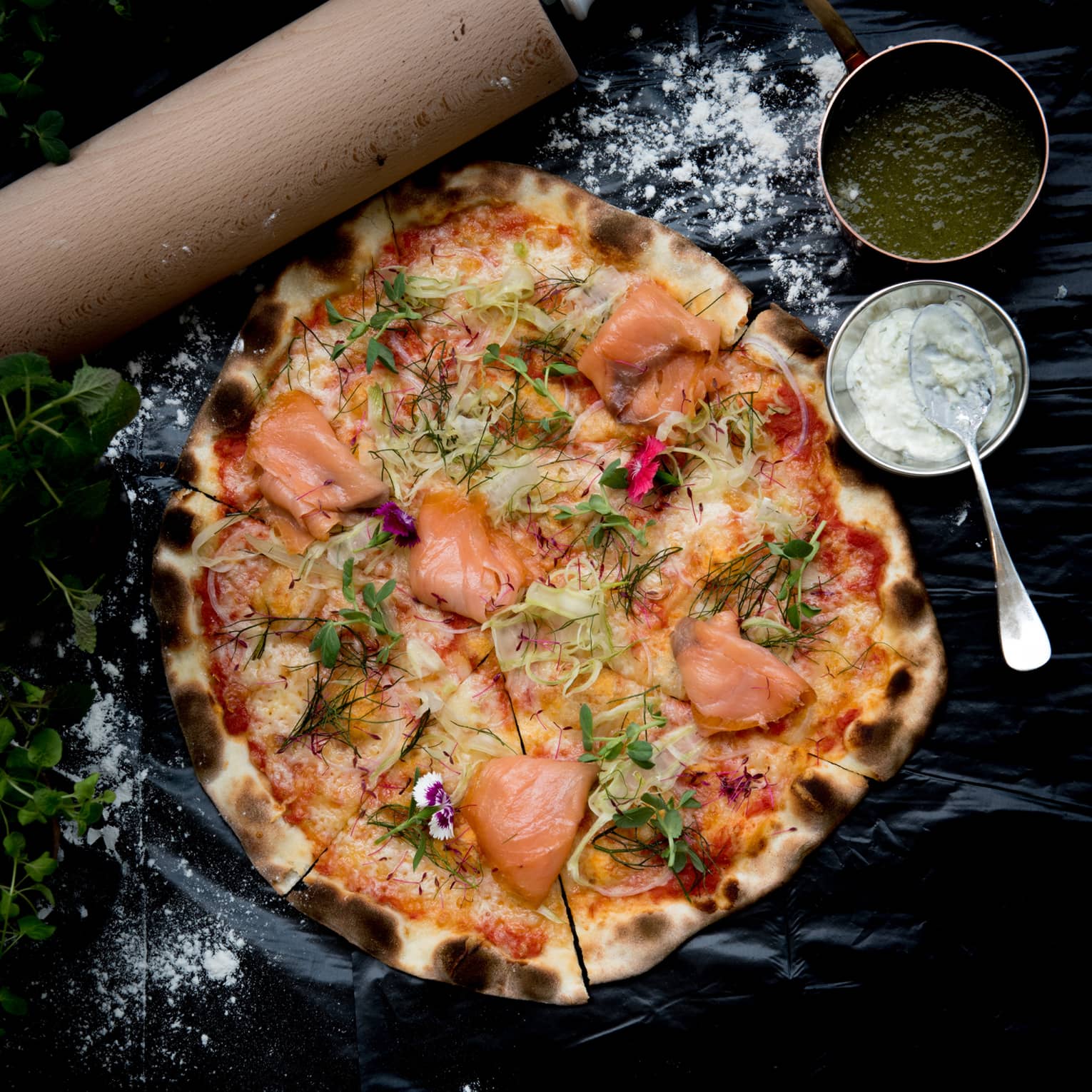 A wood-fired pizza topped with smoked salmon, fennel and dill, bowls of green and white sauces on the side. 