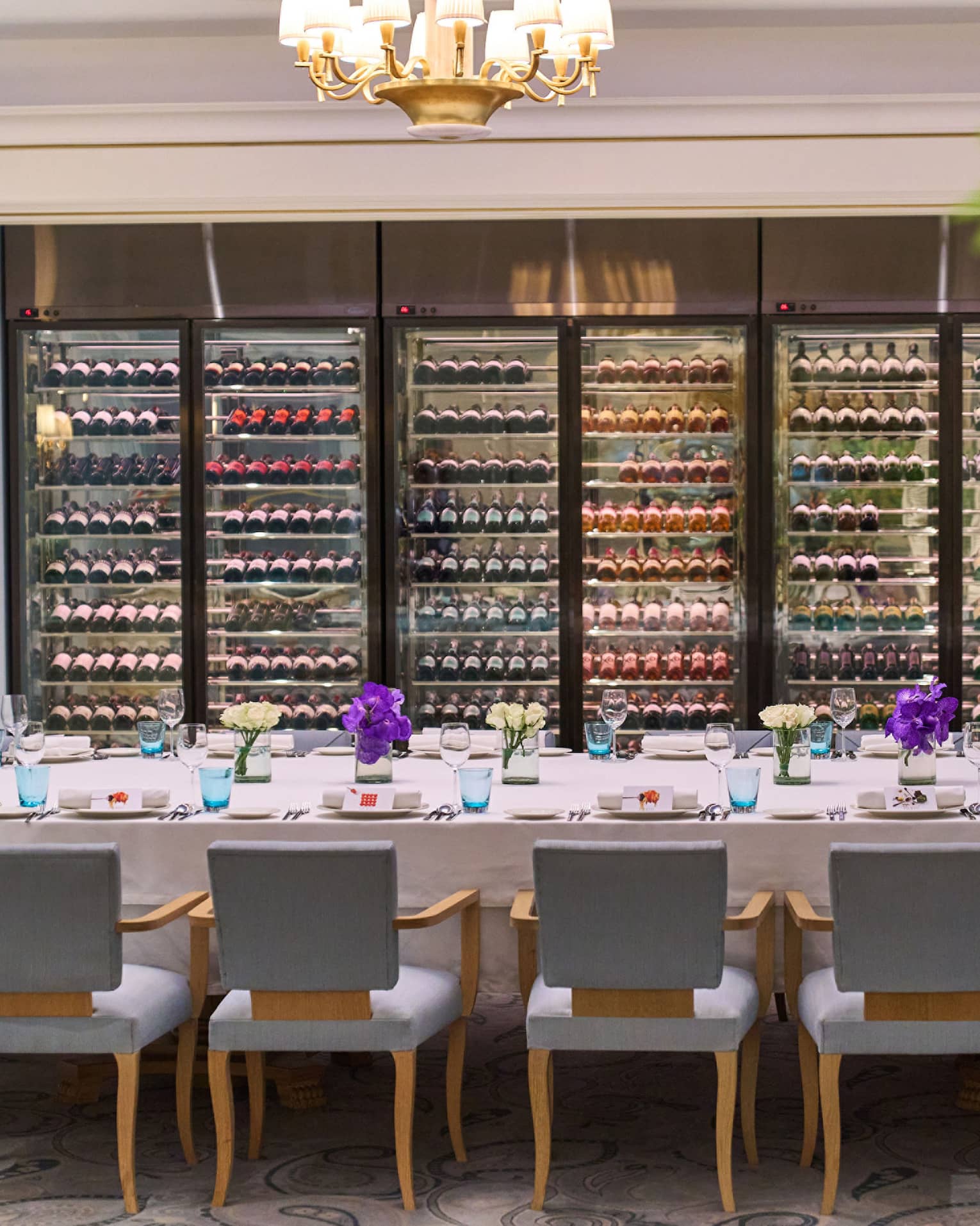 A large dining table with elegant place settings and lined by comfortable chairs before a wall of well-stocked wine fridges.