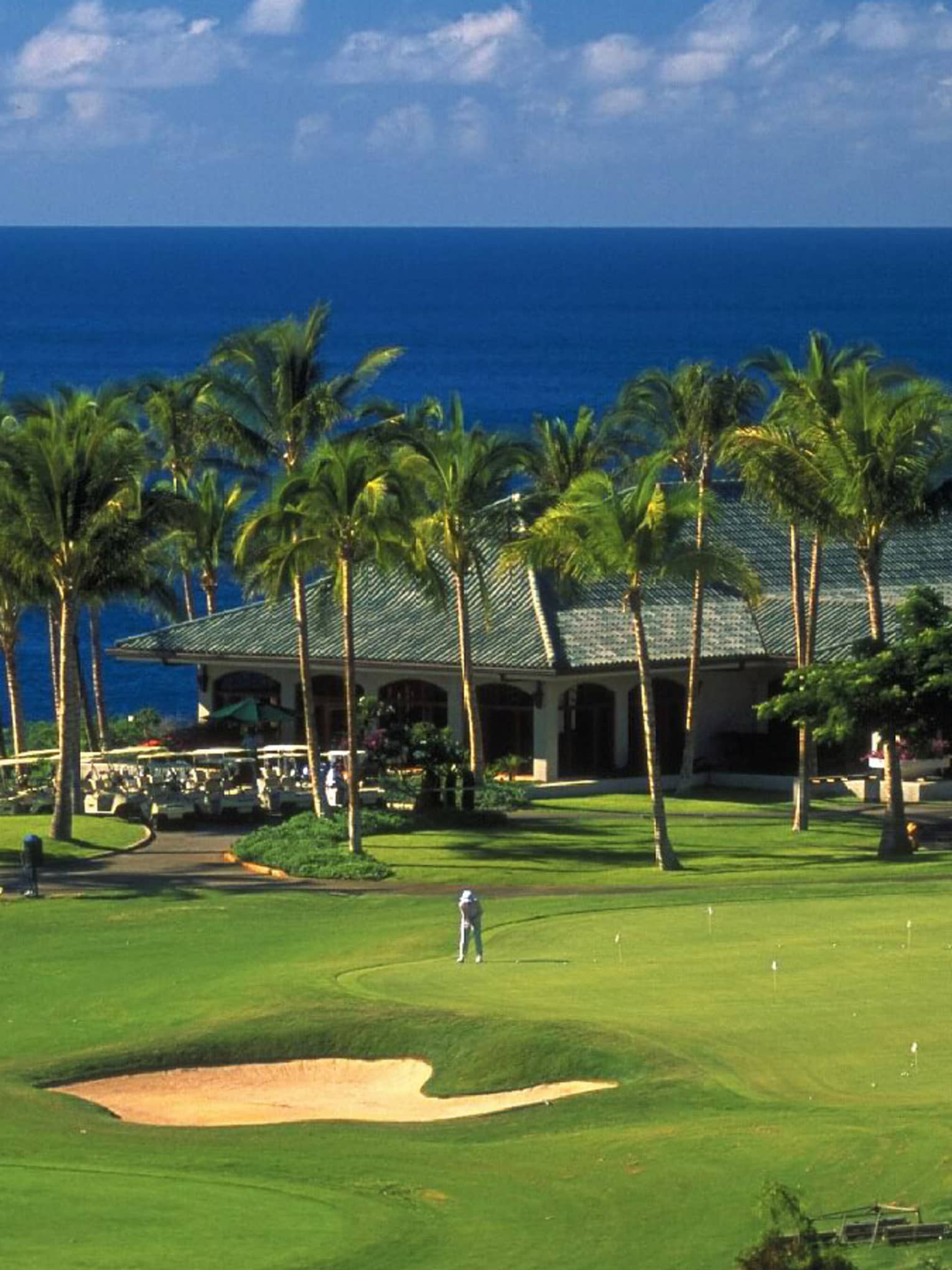 A guest plays golf on a sunny green. Palm trees are in front of a low-roofed clubhouse with the ocean in the background.