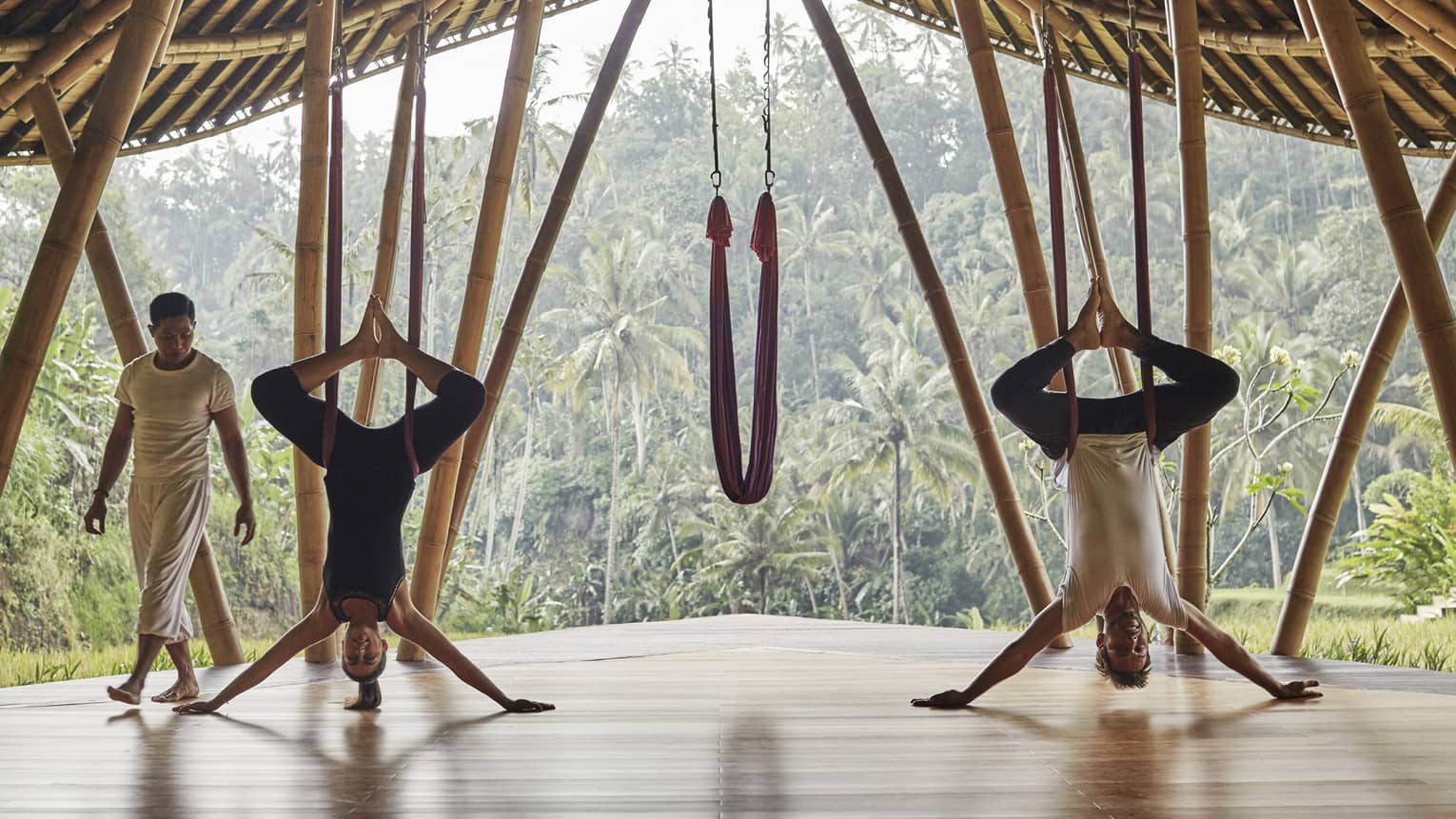 Woman and man hang upside-down on yoga hammocks under wood pavilion as instructor looks on