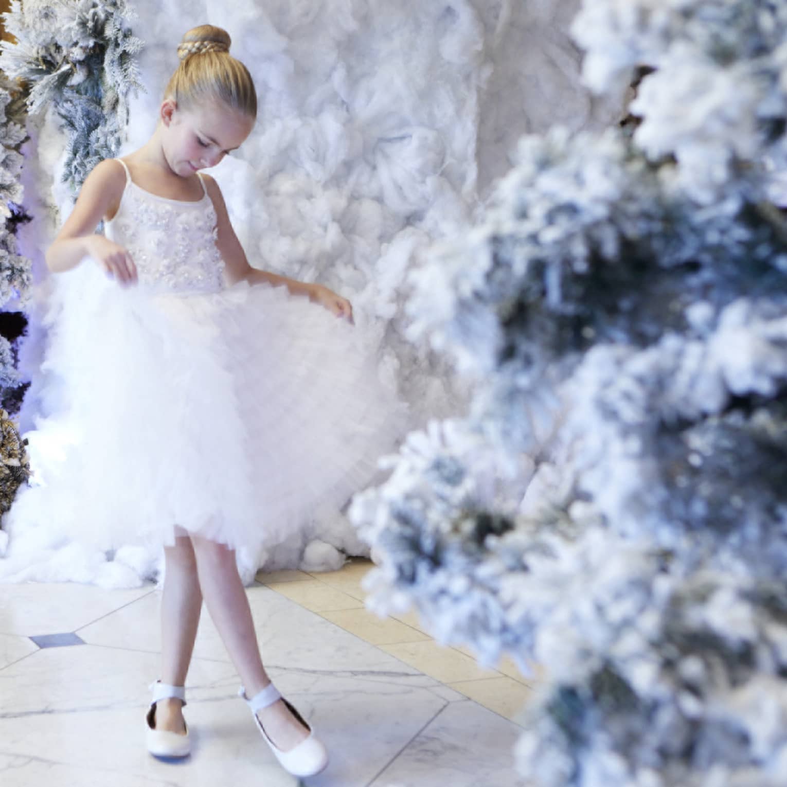 A young girl in a white ballerina dress surrounded by flowers and plants.