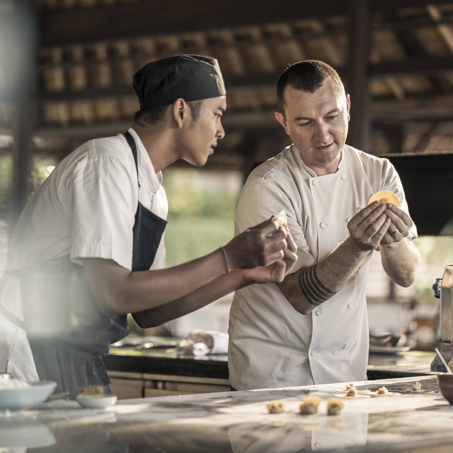 A chef teaching a student how to make pasta, folding dough with filling into the shape of a tortellini, in a sunlit kitchen.