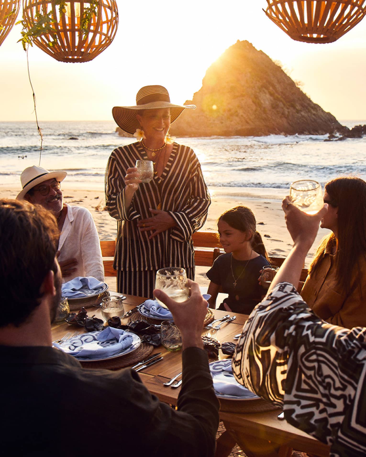 At a set table on the beach, the sun setting over the water behind them, a group of guests raise their glasses in a toast.