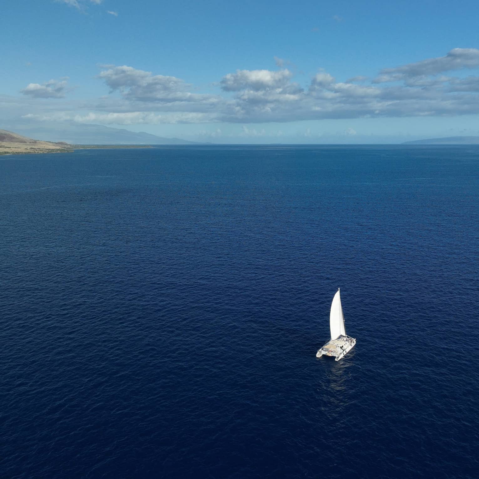 Aerial view of a catamaran as it sails through the vast, calm open waters toward the faded silhouette of distant mountains.  