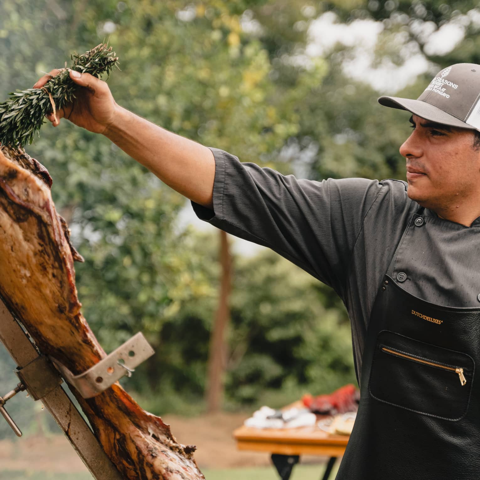 Chef wearing dark grey shirt, black apron and grey-and-white ball cap holds a bundle of herbs in one hand as he swipes it over a large piece of meat hanging on an outdoor grill