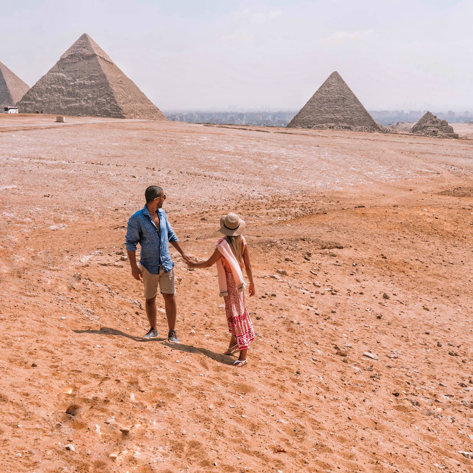 A couple holding hands turn back to look across the arid desert at the giant pyramids in the distance.