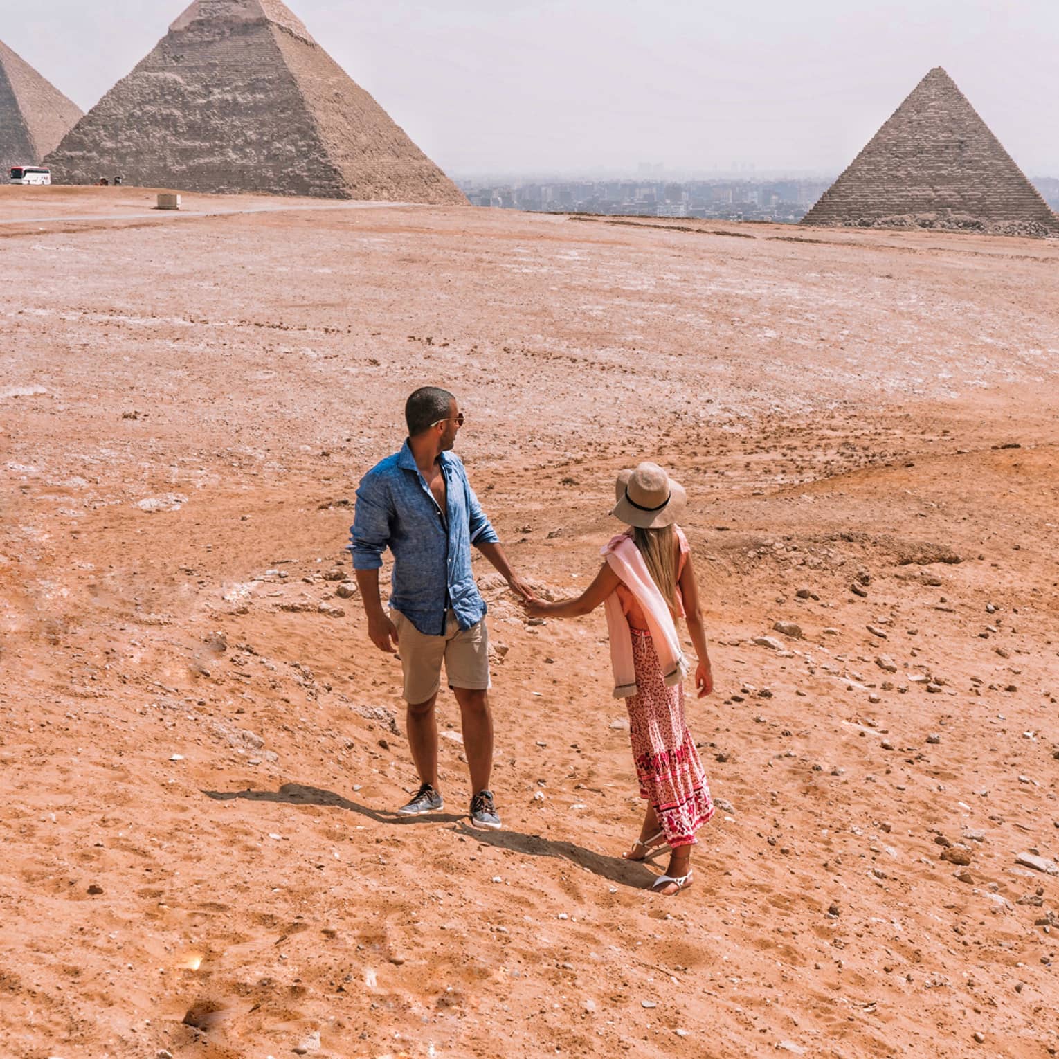 A couple holding hands turn back to look across the arid desert at the giant pyramids in the distance. 