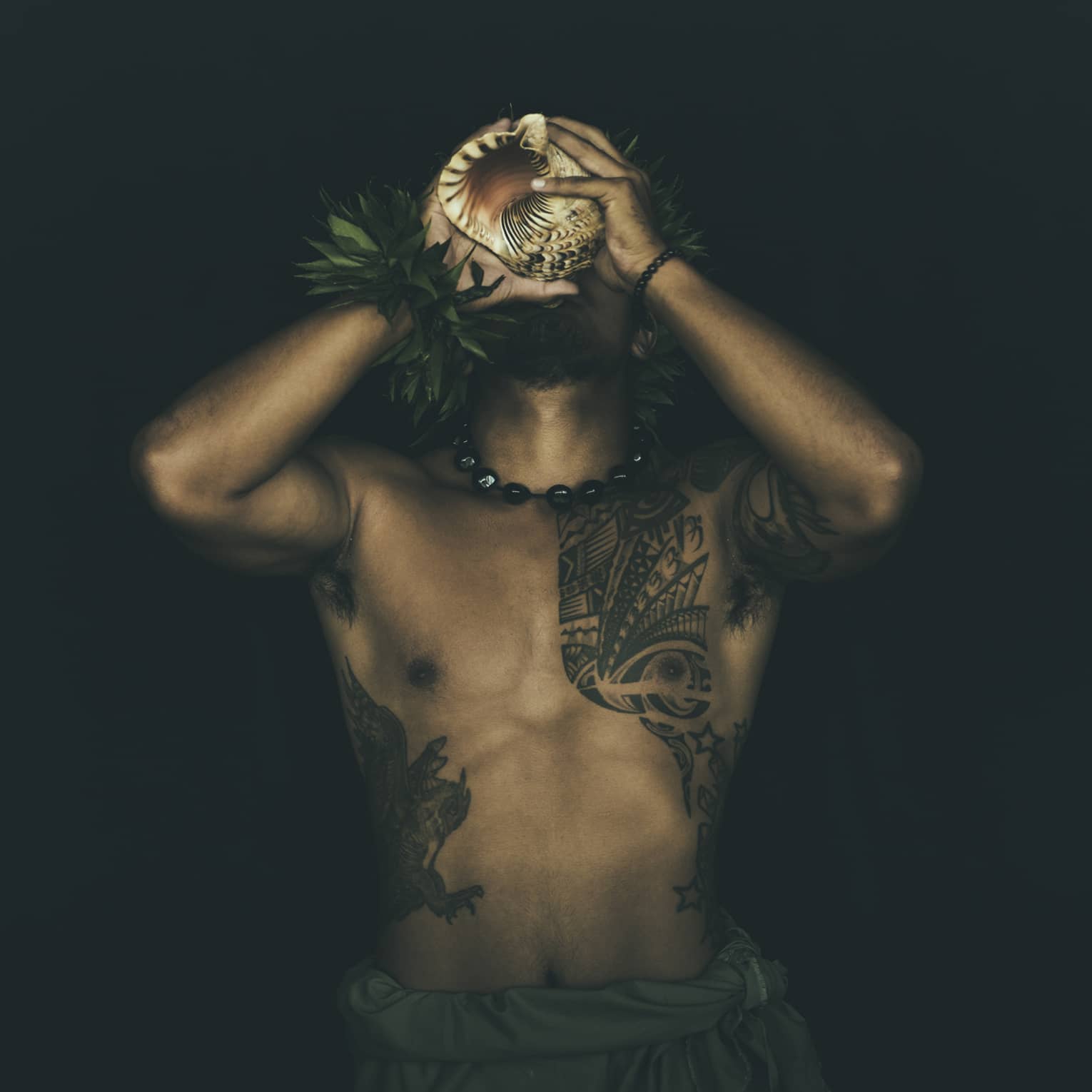 Shirtless tattooed Kānaka Maoli man plays traditional conch shell like a trumpet in the night