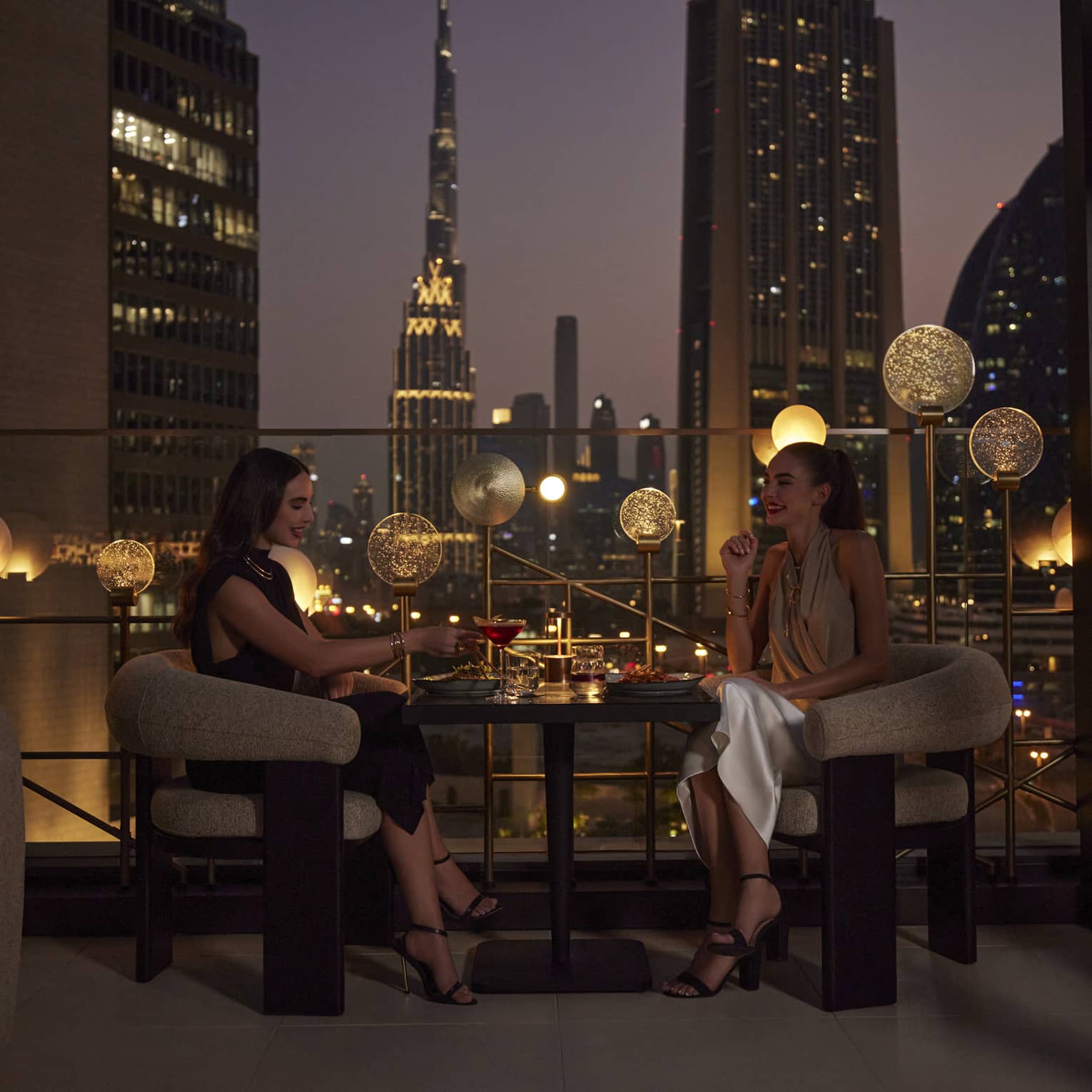 Two women sit at a cocktail table on an outdoor rooftop patio overlooking the Dubai skyline at night
