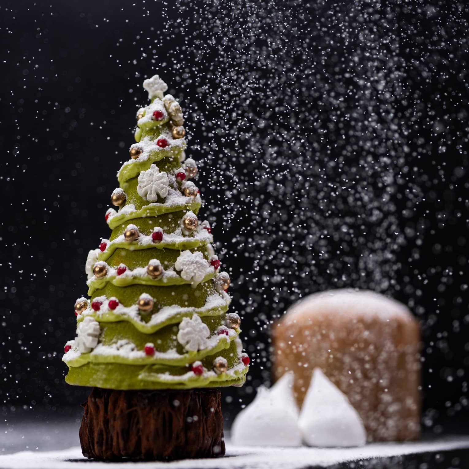 Dessert shaped like Christmas tree sits on a table as powdered sugar drops like snow from above, all on a black backdrop