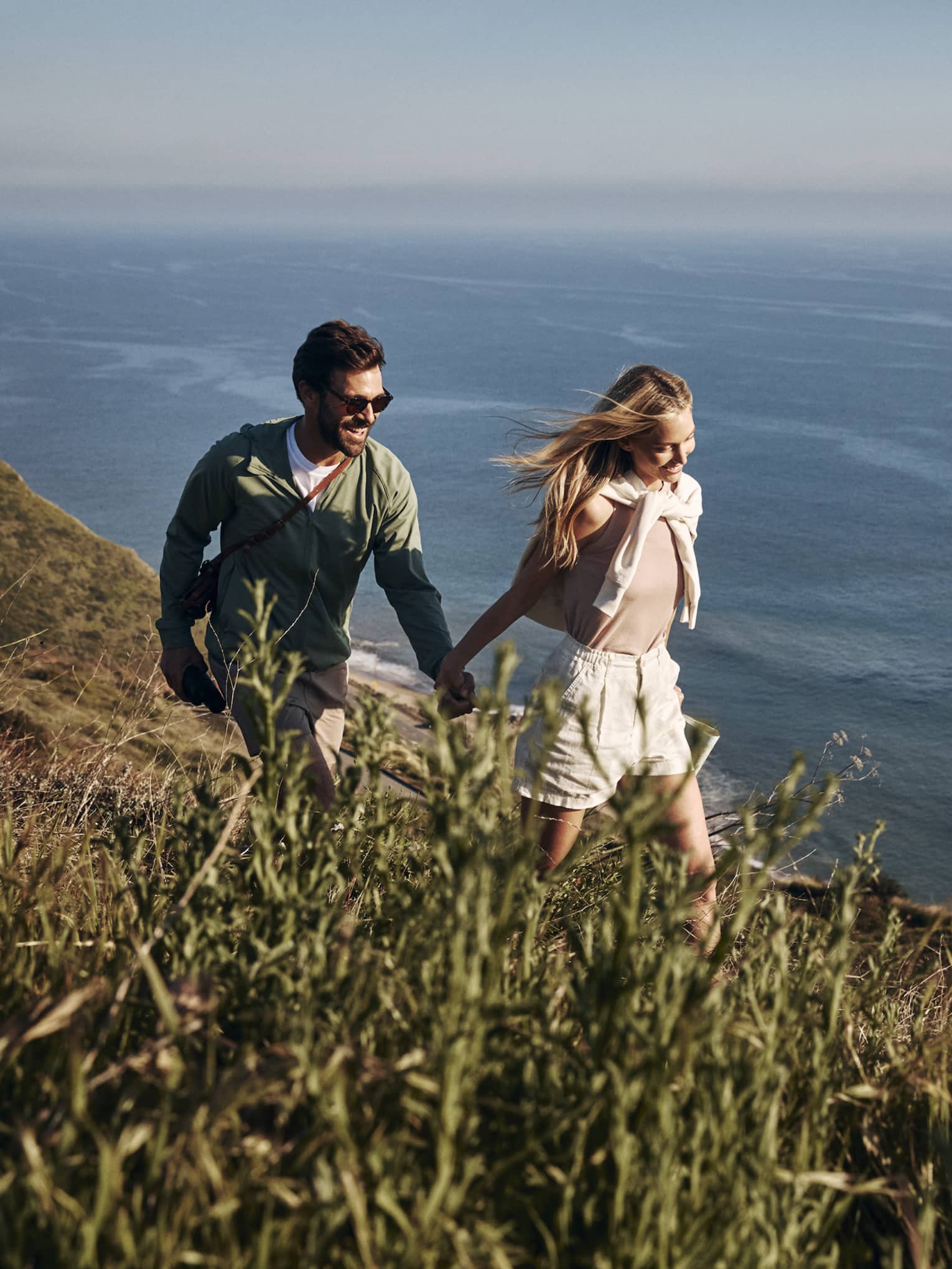 The sun shining on a smiling couple holding hands as they hike along a grassy cliff. Open water ripples below them.
