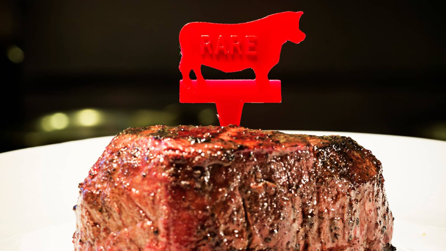 Close-up of thick filet mignon steak grilled with pepper seasoning, plastic red cow toothpick reading rare