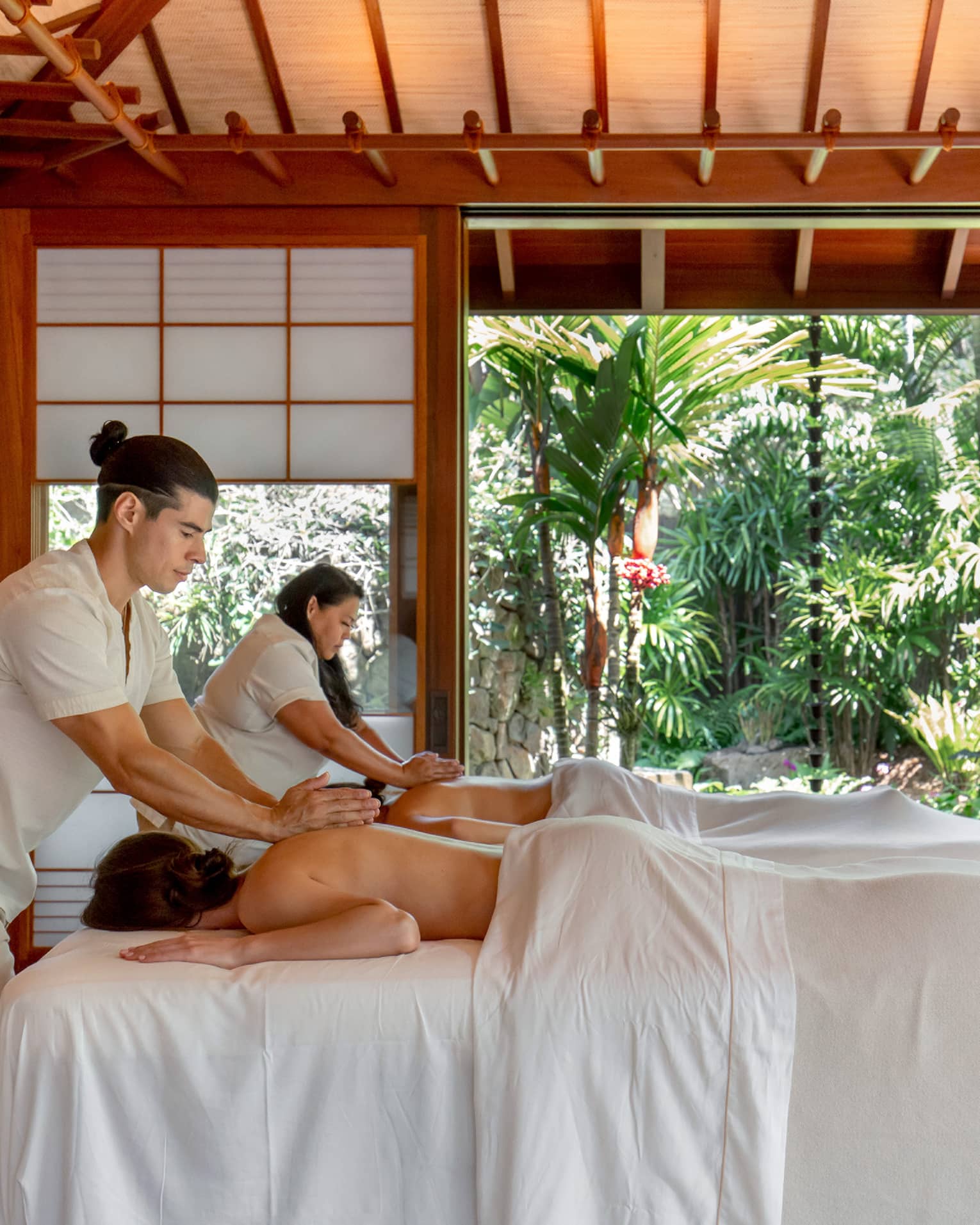 Two guests receive massages in a bright wooden hut with frosted glass windows and an entrance that opens to abundant trees.