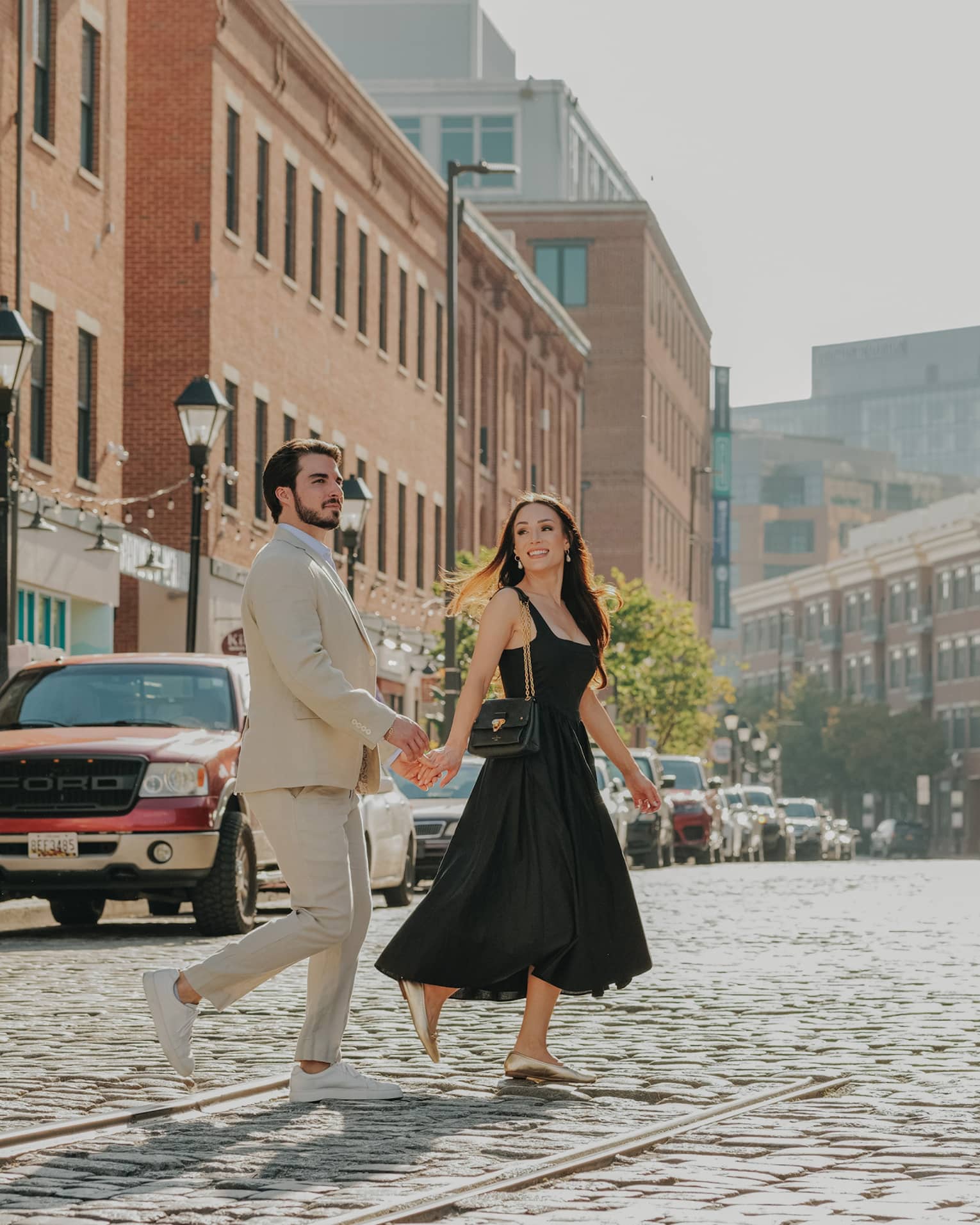 A smiling, well-dressed couple cross a cobbled street lined with stores in red-brick buildings, streetlights and parked cars.