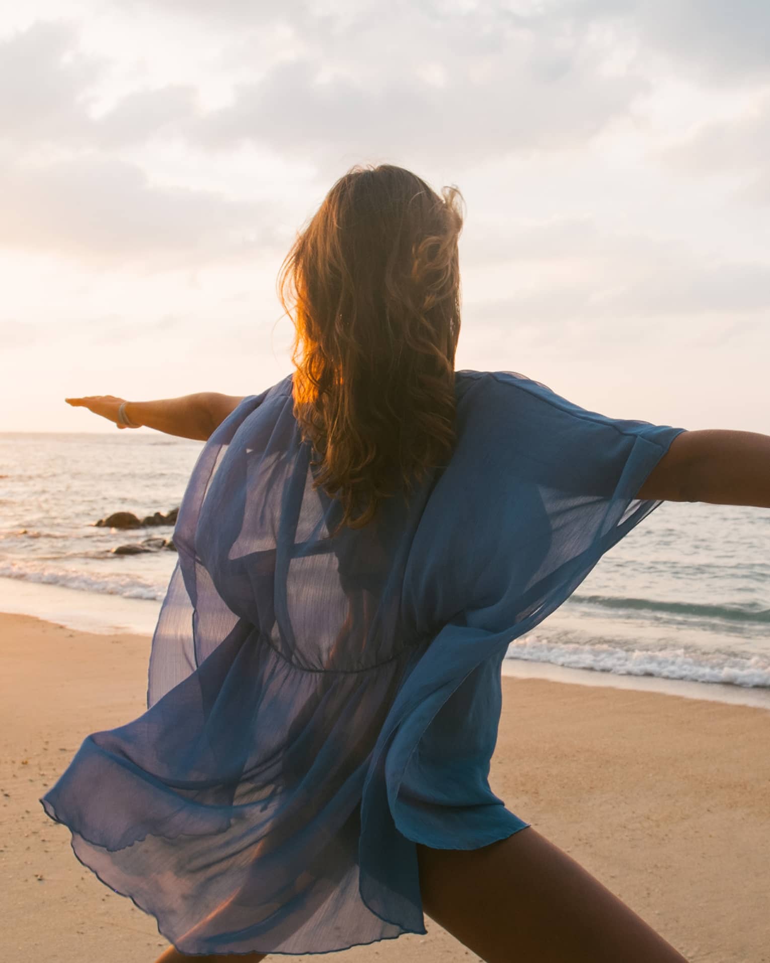 Woman wearing sheer beach cover kneels with arms outstretched in yoga pose on sandy beach by water