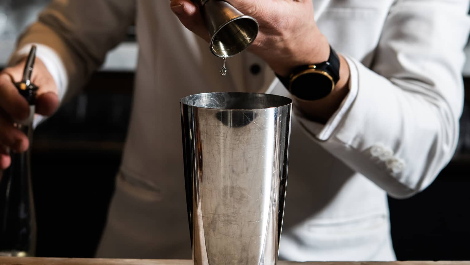 A man in an all-white suit mixes a cocktail in a silver shaker.