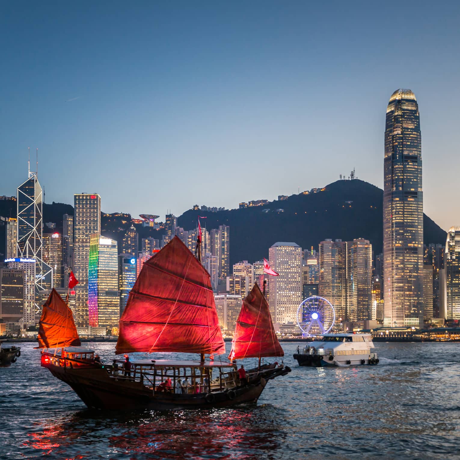 A traditional Hong Kong junk boat with red sails gliding through Victoria Harbour at dusk, with the city’s illuminated skyline in the background