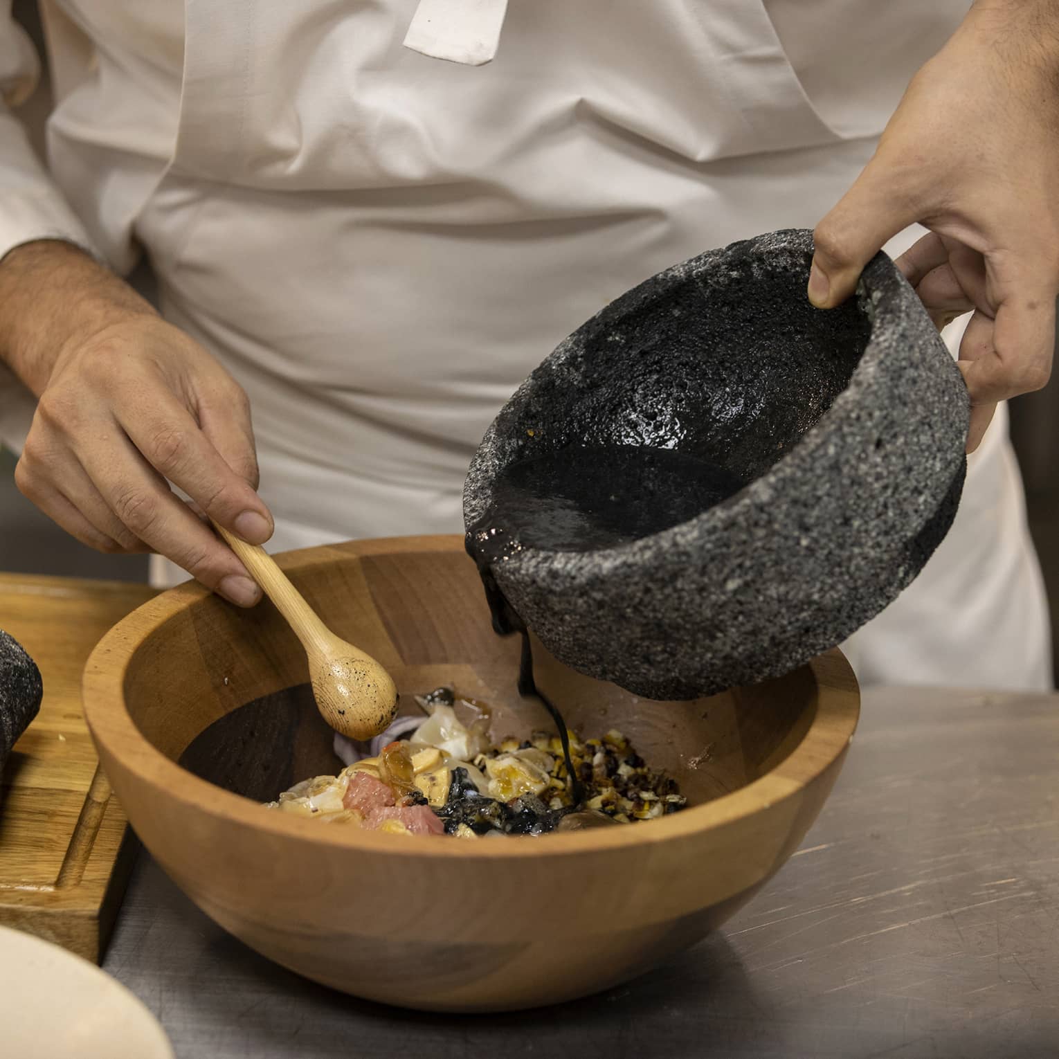 Chef pours black sauce from a stone bowl into a wood bowl of ingredients, a small wooden spoon in the other hand. 