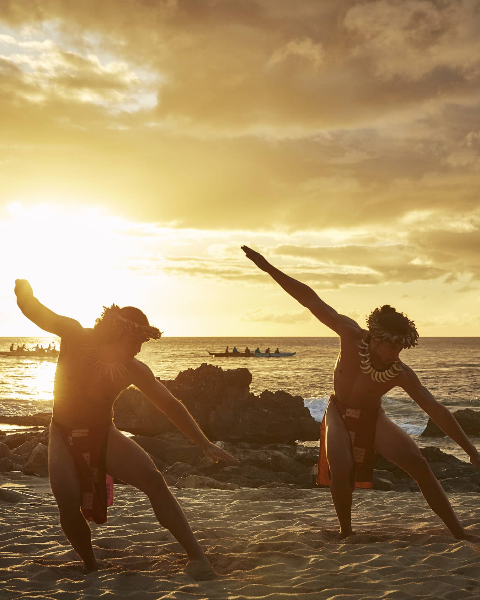 Two Hawaiian dancers with arms outstretched pose on sandy beach at sunset